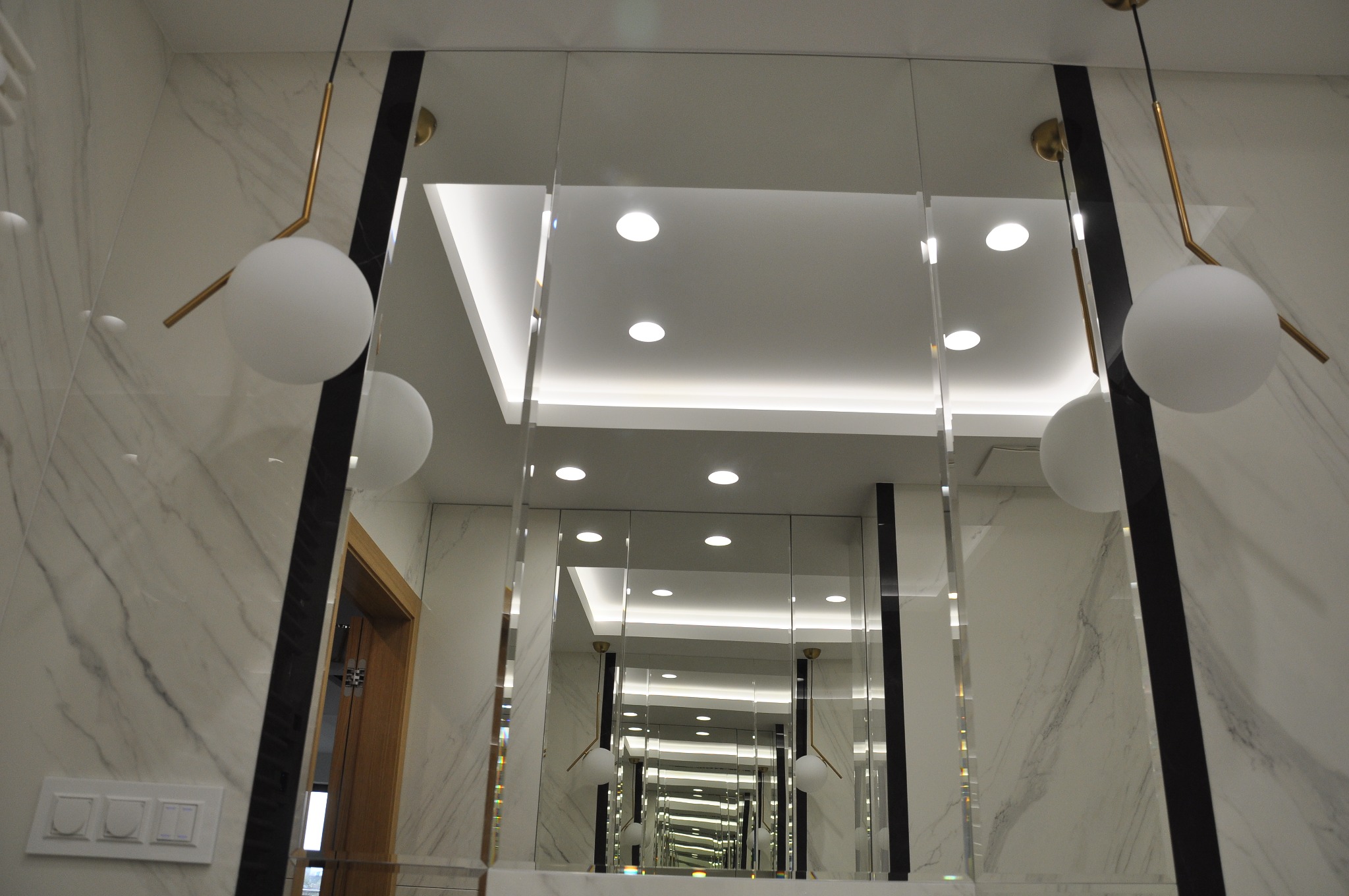 Nowoczesna łazienka z lustrem w długim, wąskim panelu, odbijającym serię identycznych paneli i lamp kulistych, marmurowe ściany, widoczny fragment drewnianych drzwi.