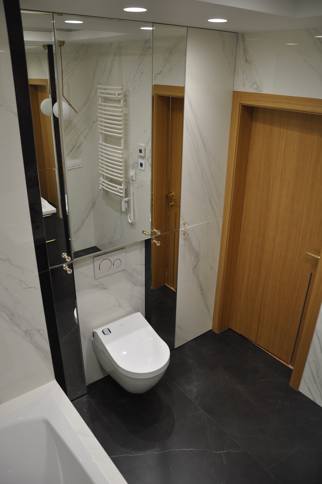 Nowoczesna łazienka z marmurowymi ścianami i czarną podłogą, wisząca toaleta z panelem sterowania, lustro z kryształowymi gałkami, drewniane drzwi.