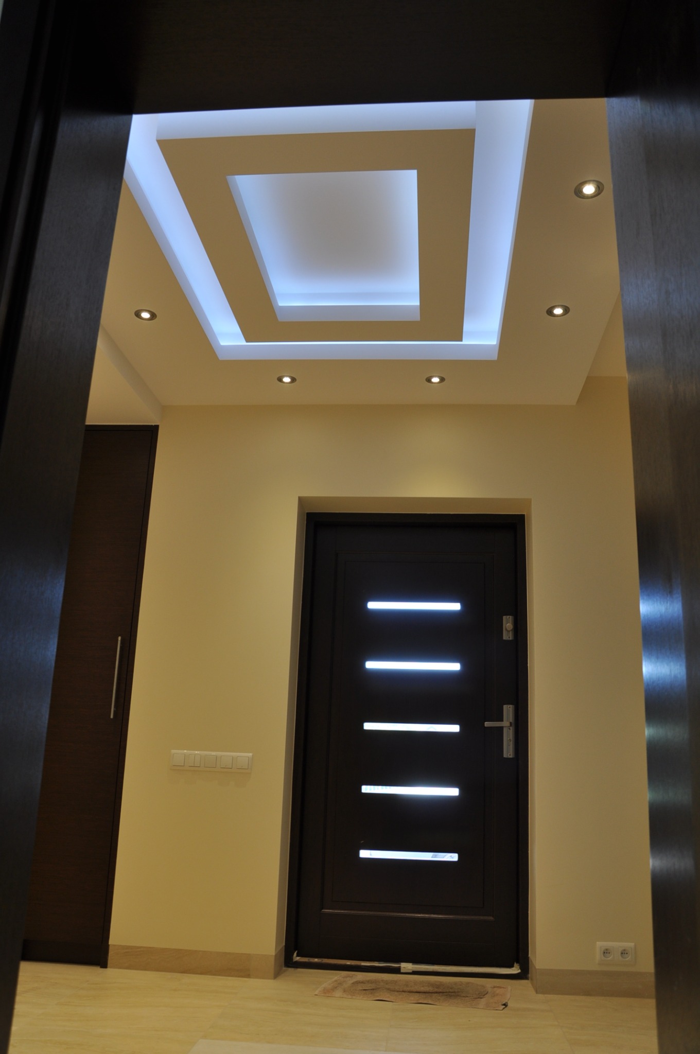 Nowoczesne wnętrze przedpokoju z podwieszanym sufitem z oświetleniem LED, ciemne drzwi wejściowe z poziomymi przeszkleniami, szafa w zabudowie i beżowe ściany.