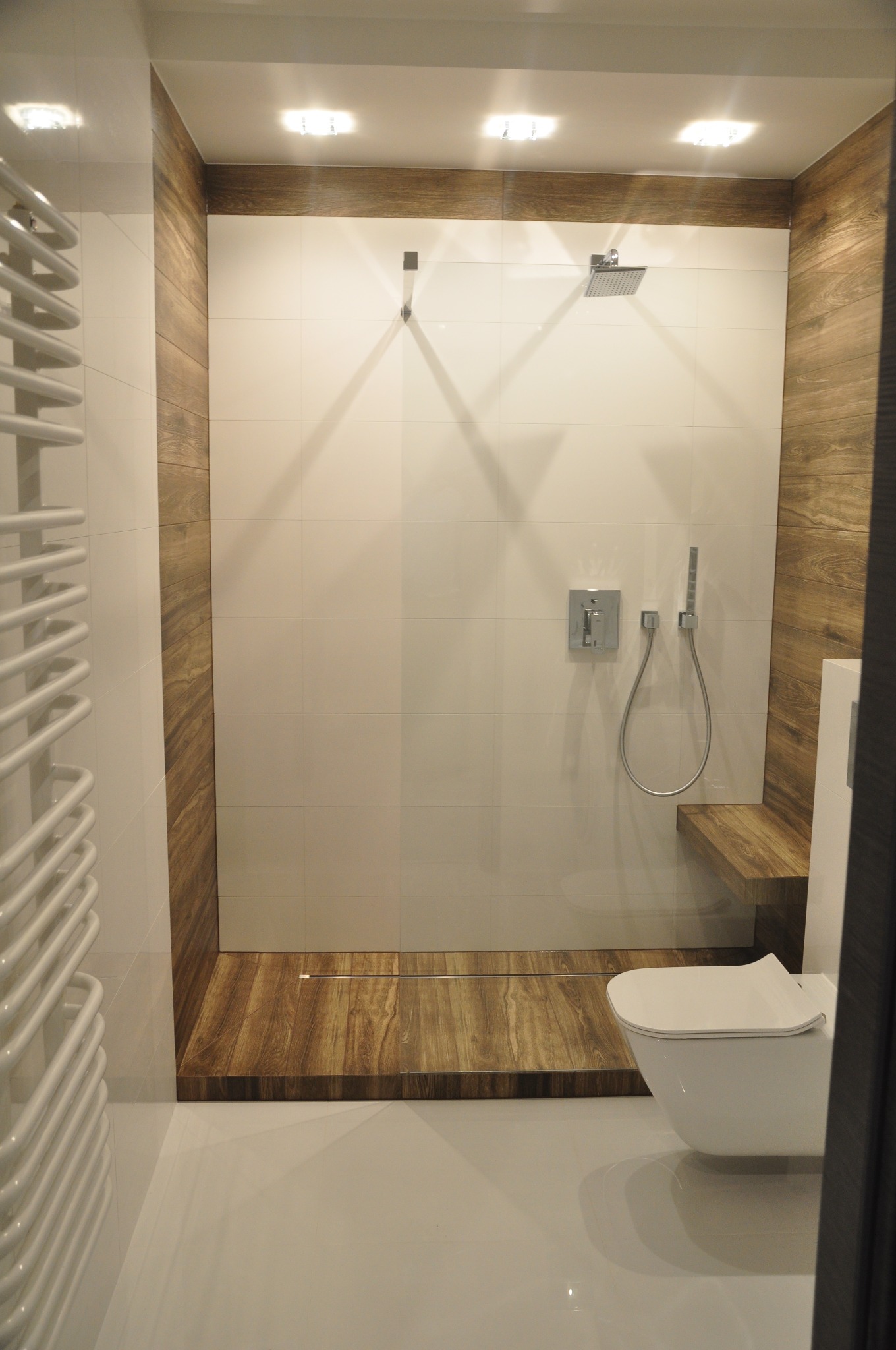 Nowoczesna kabina prysznicowa walk-in z drewnianym wykończeniem, białe płytki ścienne, deszczownica i bateria podtynkowa, toaleta wisząca z deską wolnoopadającą, grzejnik drabinkowy.