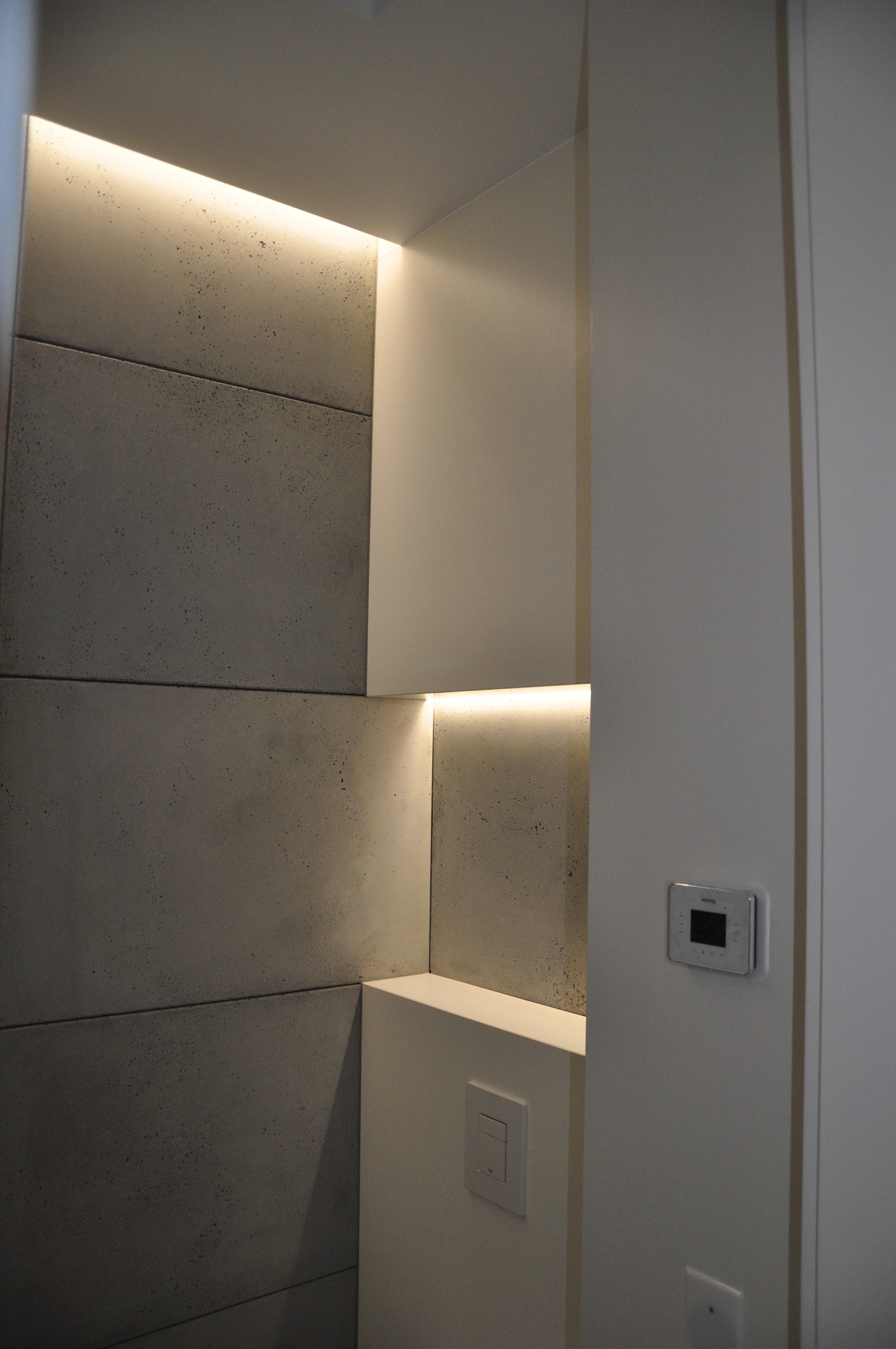Wnęka z szarymi panelami imitującymi beton, podświetlona ciepłym światłem LED, z widocznym panelem sterowania na ścianie.