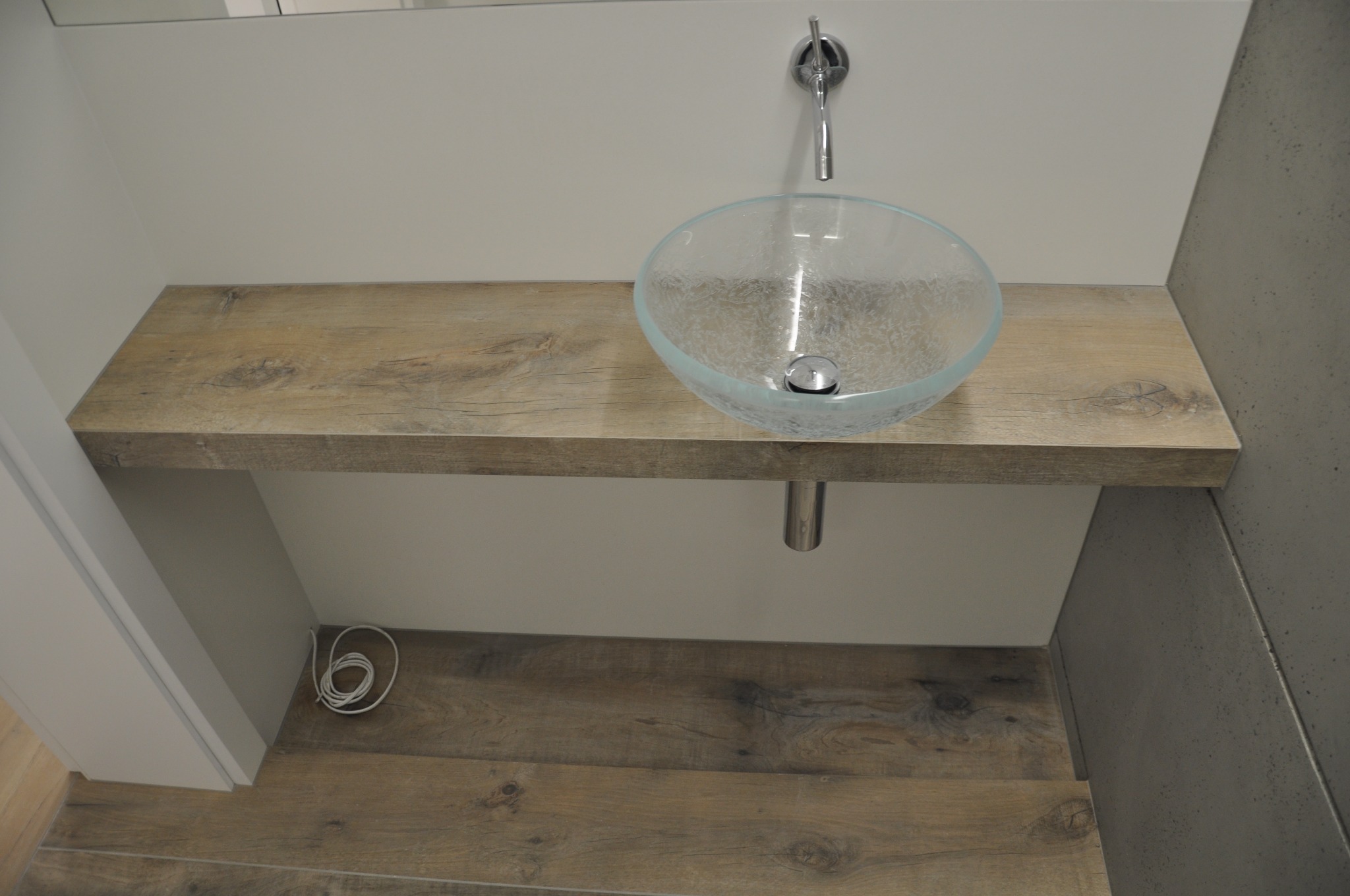 Szklana umywalka nablatowa zamontowana na drewnianym blacie w nowoczesnej łazience, widoczne przewody pod umywalką.