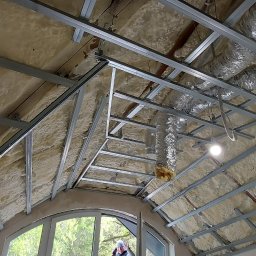 Bohdan Portsina - Montaż metalowej konstrukcji pod izolację poddasza z widocznymi przewodami wentylacyjnymi i oknem dachowym w tle.