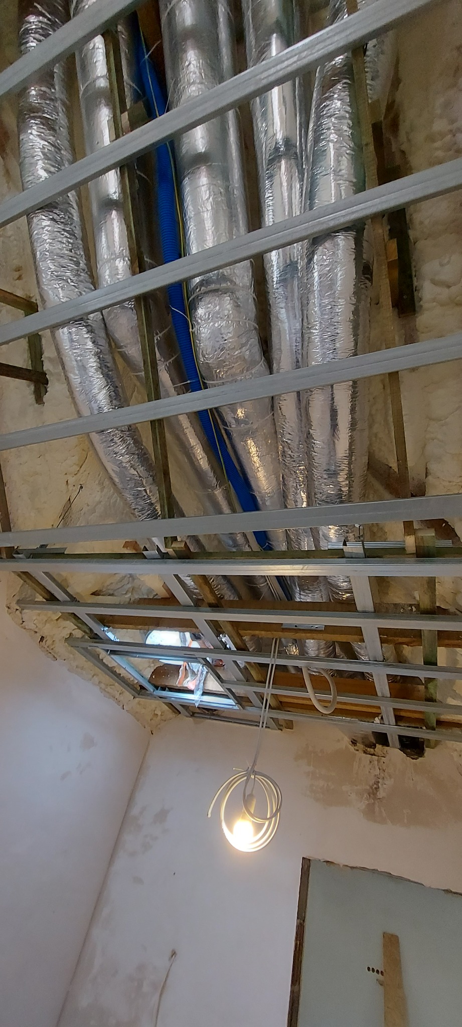 Montaż instalacji wentylacyjnej w przestrzeni pod przyszłym sufitem, widoczne rury w izolacji, metalowy stelaż i fragment ściany z widocznymi śladami po pracach remontowych.