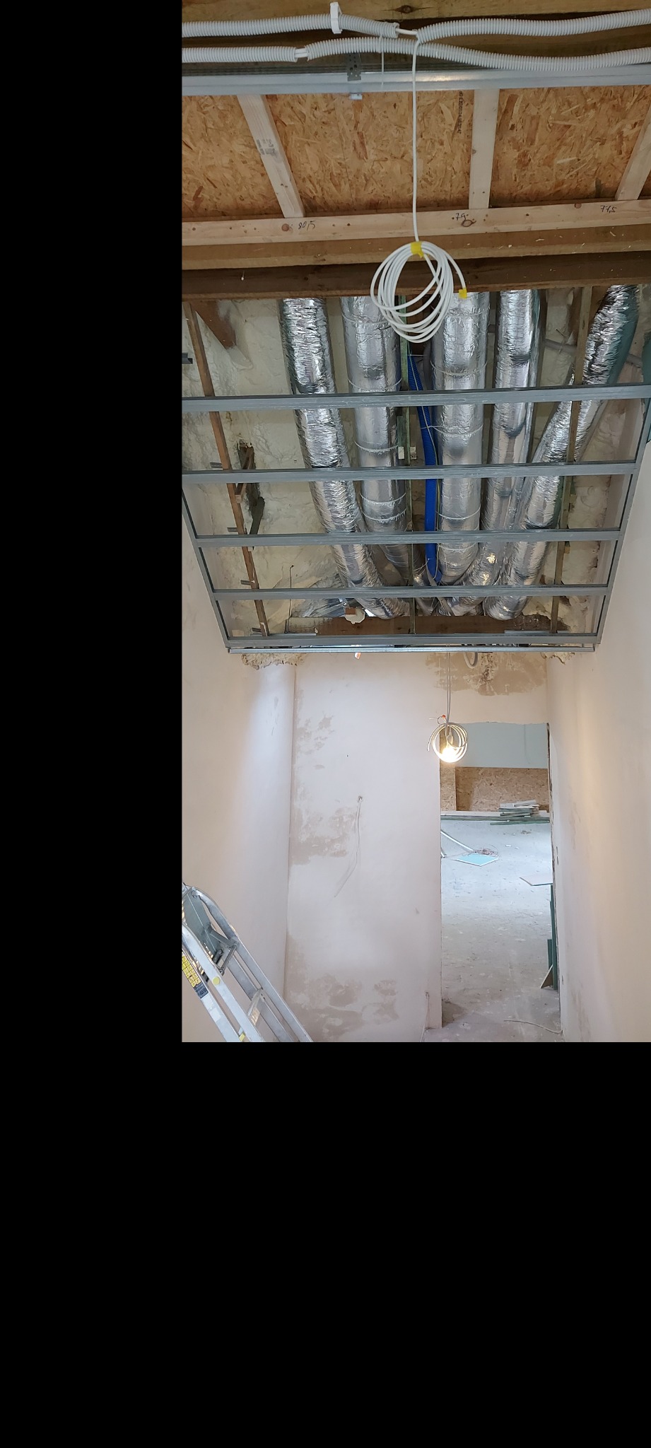 Montaż instalacji wentylacyjnej w przestrzeni pod sufitem, widoczne rury wentylacyjne w izolacji oraz metalowa konstrukcja nośna, w tle surowa ściana i drabina.