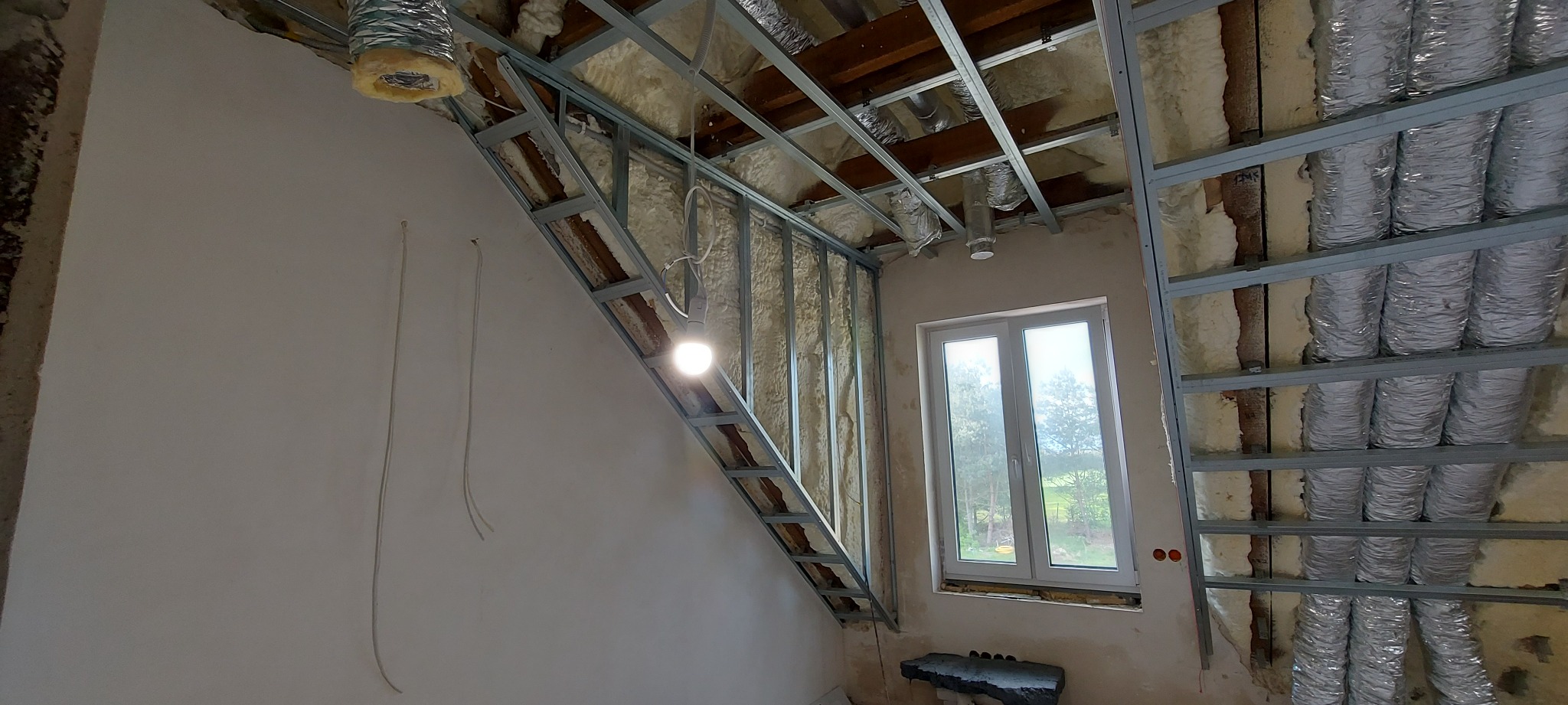 Szkielet metalowy konstrukcji sufitu podwieszanego, wypełniony wełną mineralną, widoczne przewody wentylacyjne, okno w tle, surowy stan wykończenia wnętrza.