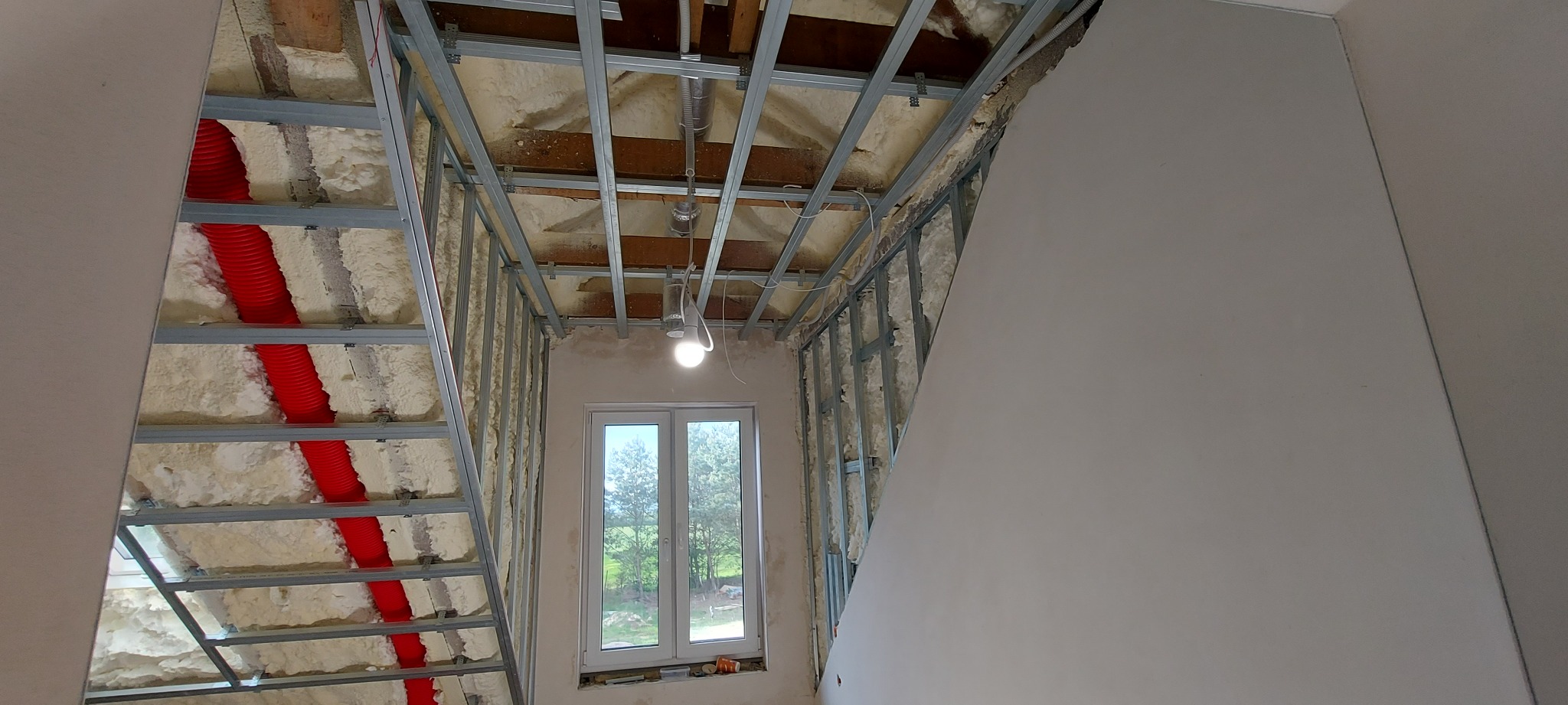 Szkielet konstrukcji sufitu i ścian z profili metalowych wypełniony pianką izolacyjną, widoczne czerwone rury wentylacyjne i okno.