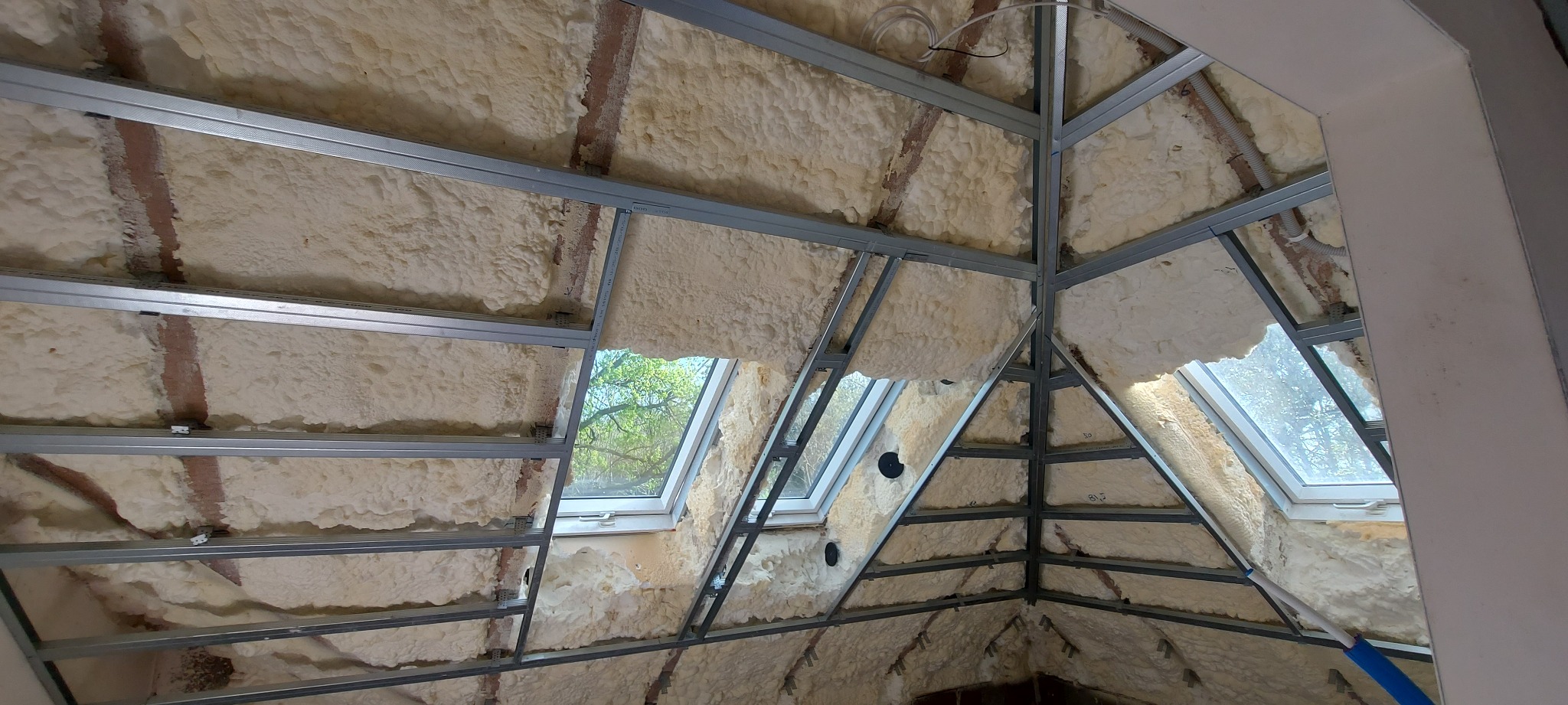 Skośny strych po ociepleniu pianką poliuretanową, widoczne profile metalowe pod konstrukcję sufitu i okna dachowe z widokiem na drzewa.