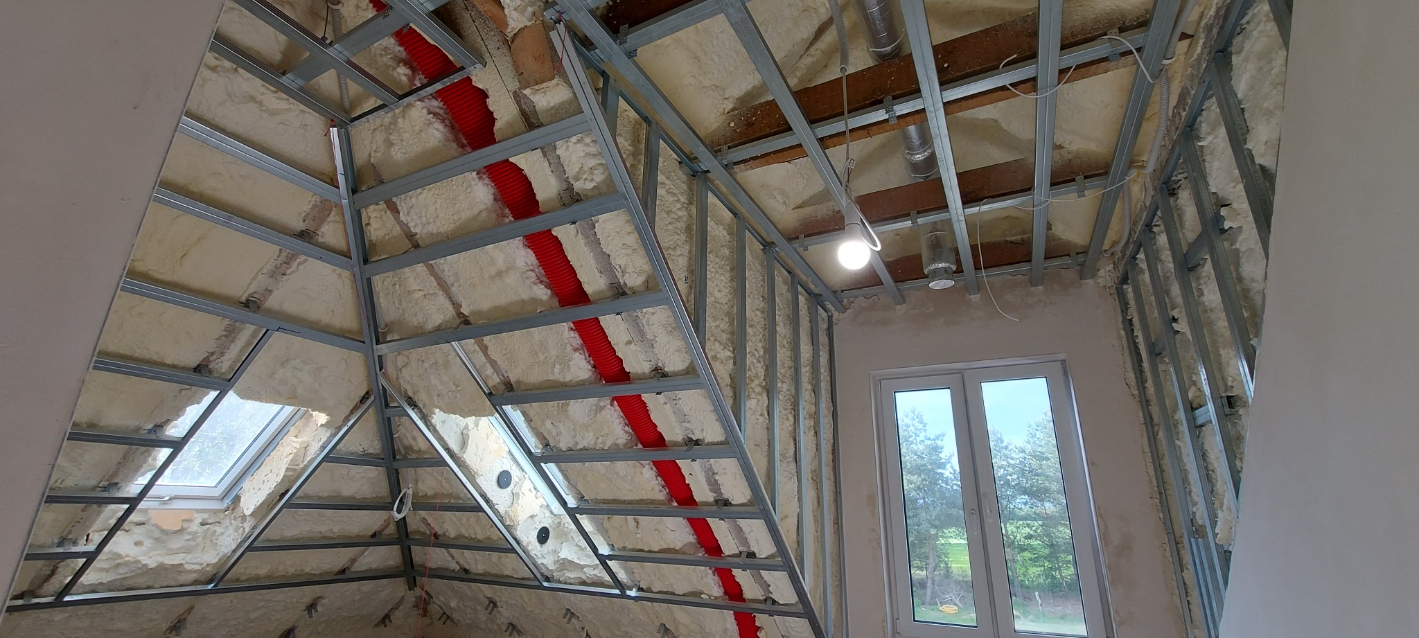 Poddasze w trakcie adaptacji: widoczne metalowe profile konstrukcyjne, ocieplenie pianą, czerwone rury instalacyjne biegnące wzdłuż krokwi, okno dachowe i okno pionowe z widokiem na zieleń.