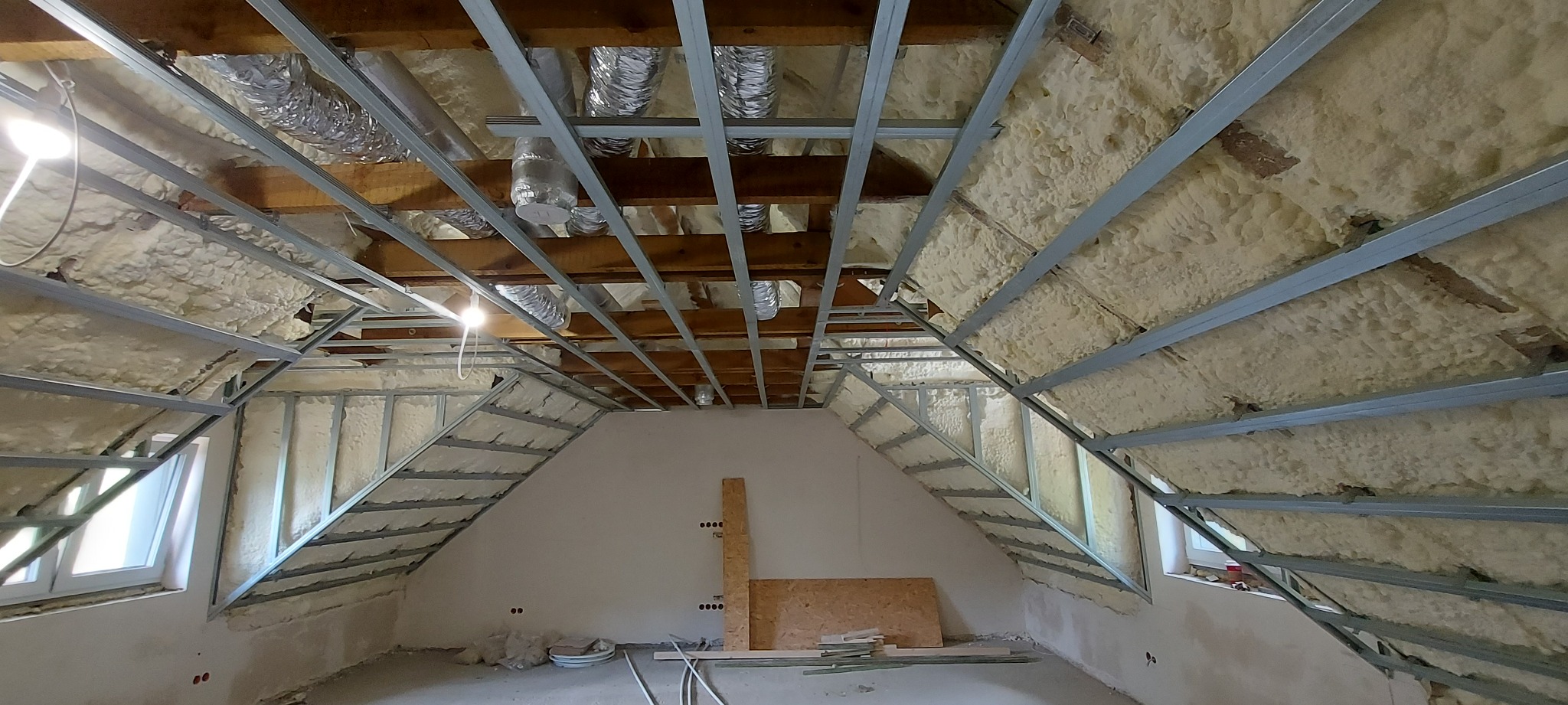 Poddasze w trakcie izolacji pianą PUR, widoczne metalowe profile konstrukcyjne, drewniane belki stropowe i fragmenty instalacji wentylacyjnej.