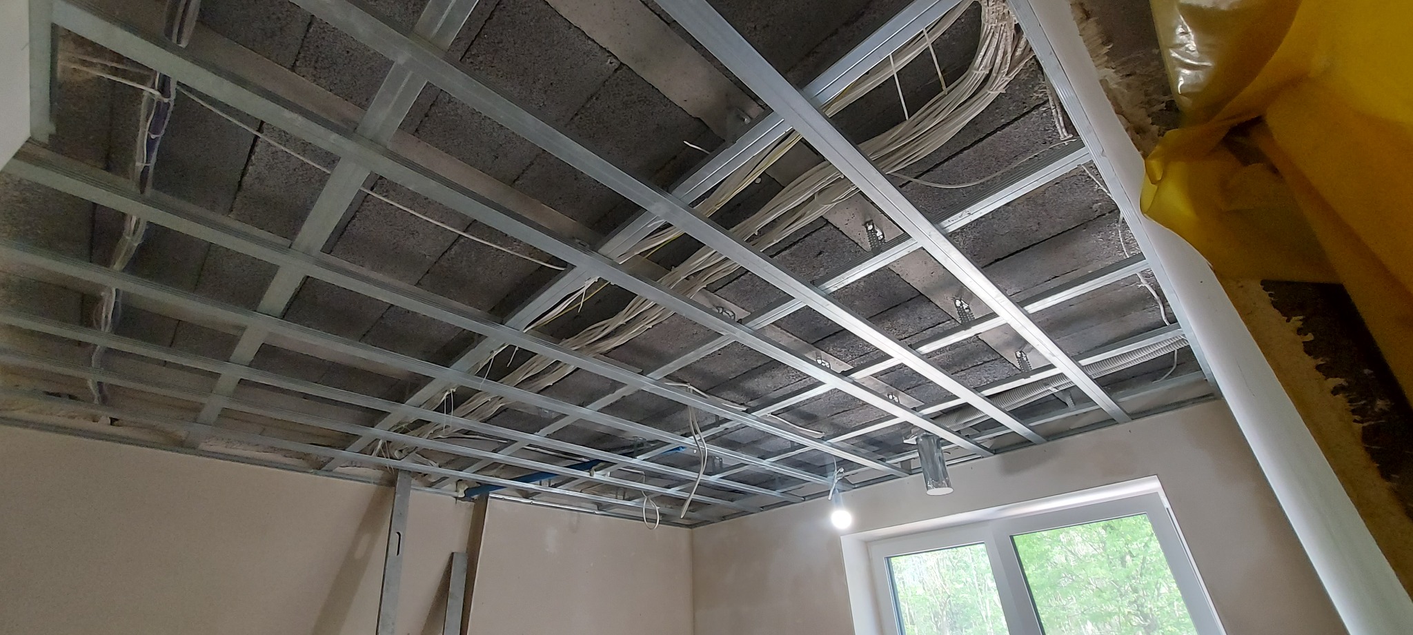 Metalowa konstrukcja sufitu podwieszanego z widocznymi kablami elektrycznymi, oświetleniem i fragmentem okna w nowym budynku.