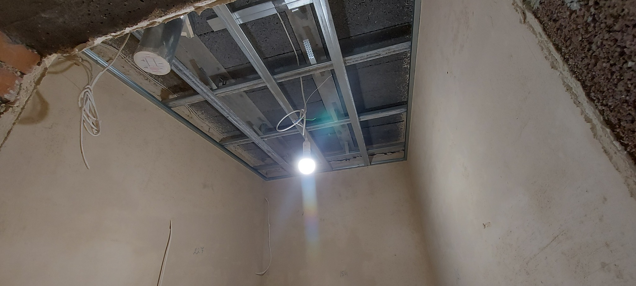 Montaż metalowej konstrukcji sufitu podwieszanego z widocznymi przewodami i żarówką tymczasową w surowym wnętrzu.