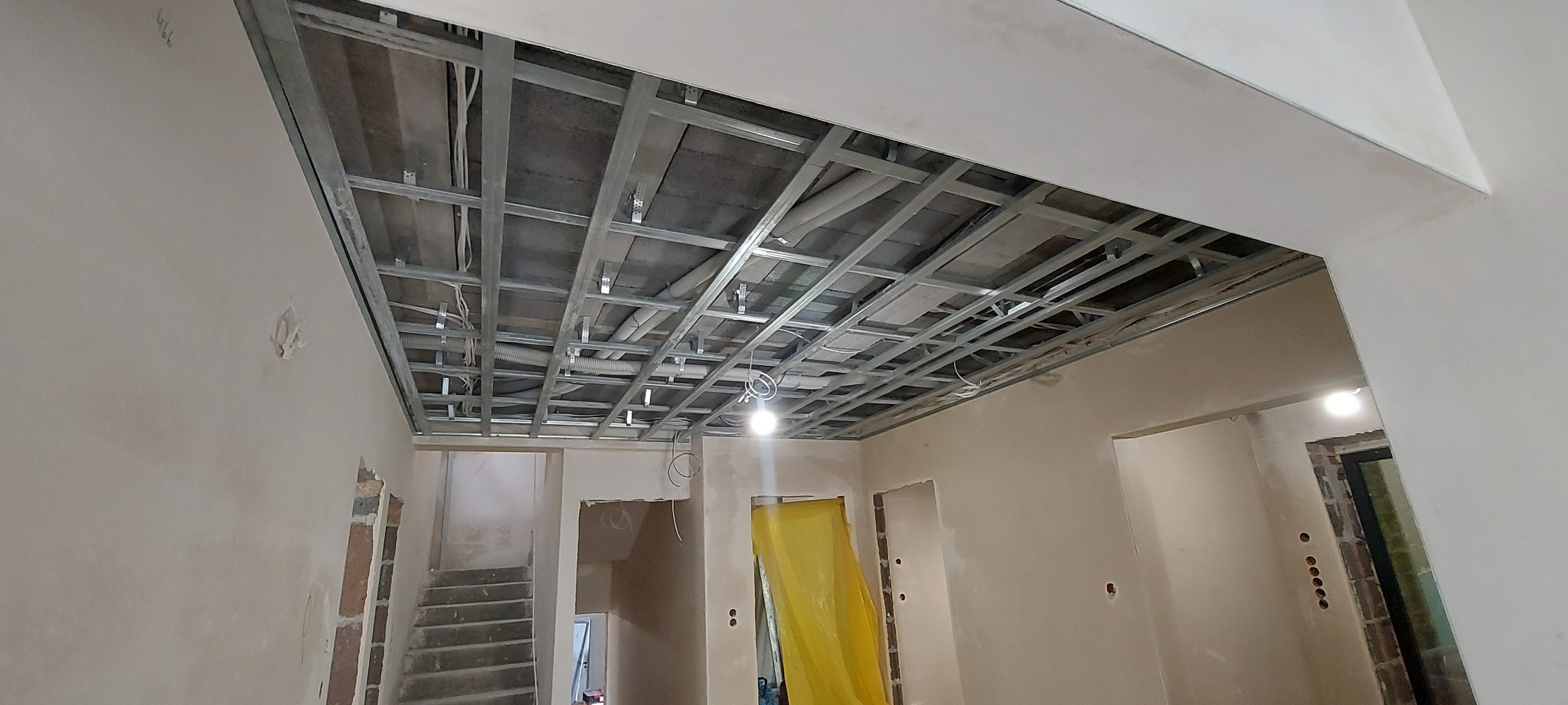 Stelaż sufitowy z profili metalowych, częściowo zakryty płytami, widoczne przewody i rury wentylacyjne, surowe ściany w tle.