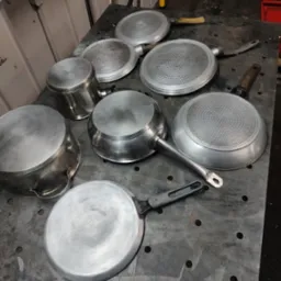 Metalowe garnki i patelnie po procesie piaskowania, leżące na perforowanym stole warsztatowym, widoczne ślady obróbki.