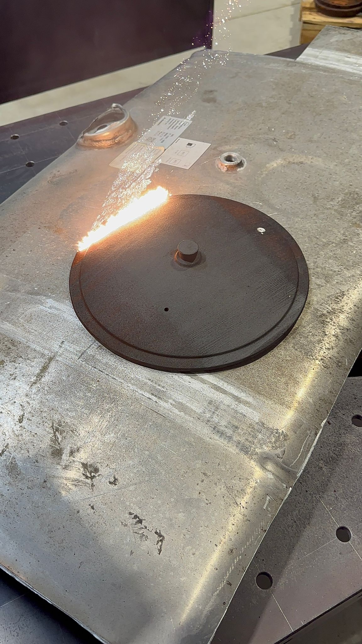 Metalowa pokrywa zbiornika po obróbce strumieniowo-ściernej, z widocznymi iskrami i pozostałościami po procesie, umieszczona na metalowej powierzchni roboczej.