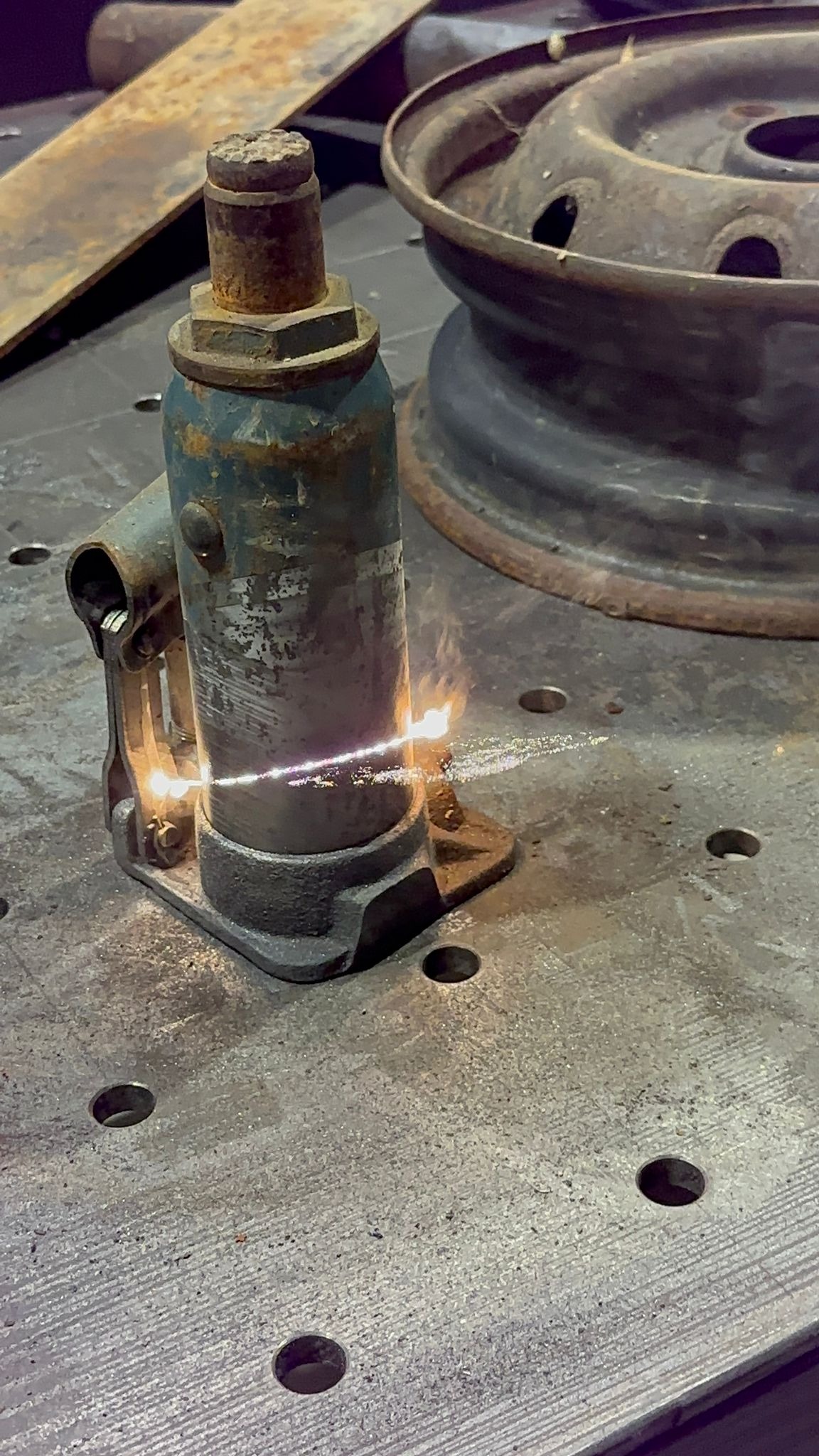 Stary, zardzewiały podnośnik hydrauliczny oczyszczany strumieniem ognia na metalowym stole warsztatowym, obok leży zardzewiała felga samochodowa.