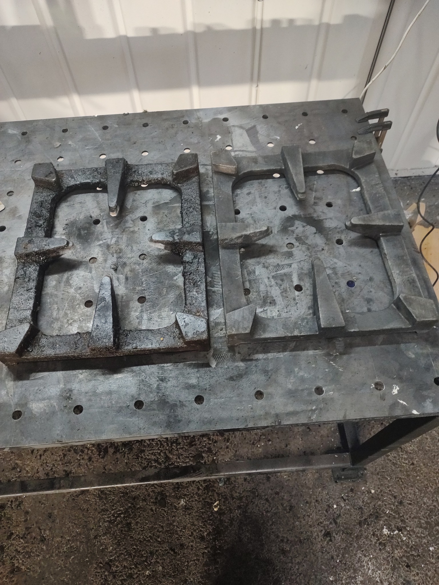 Dwie metalowe formy odlewnicze, jedna z widocznymi śladami korozji i zabrudzeń, druga czystsza, leżące na perforowanym stole warsztatowym w hali produkcyjnej.