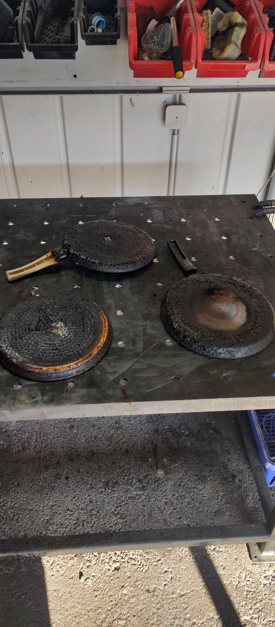 Trzy mocno zabrudzone i przypalone elementy kuchenne, prawdopodobnie płyty grzewcze, leżące na perforowanym metalowym stole warsztatowym w warsztacie.