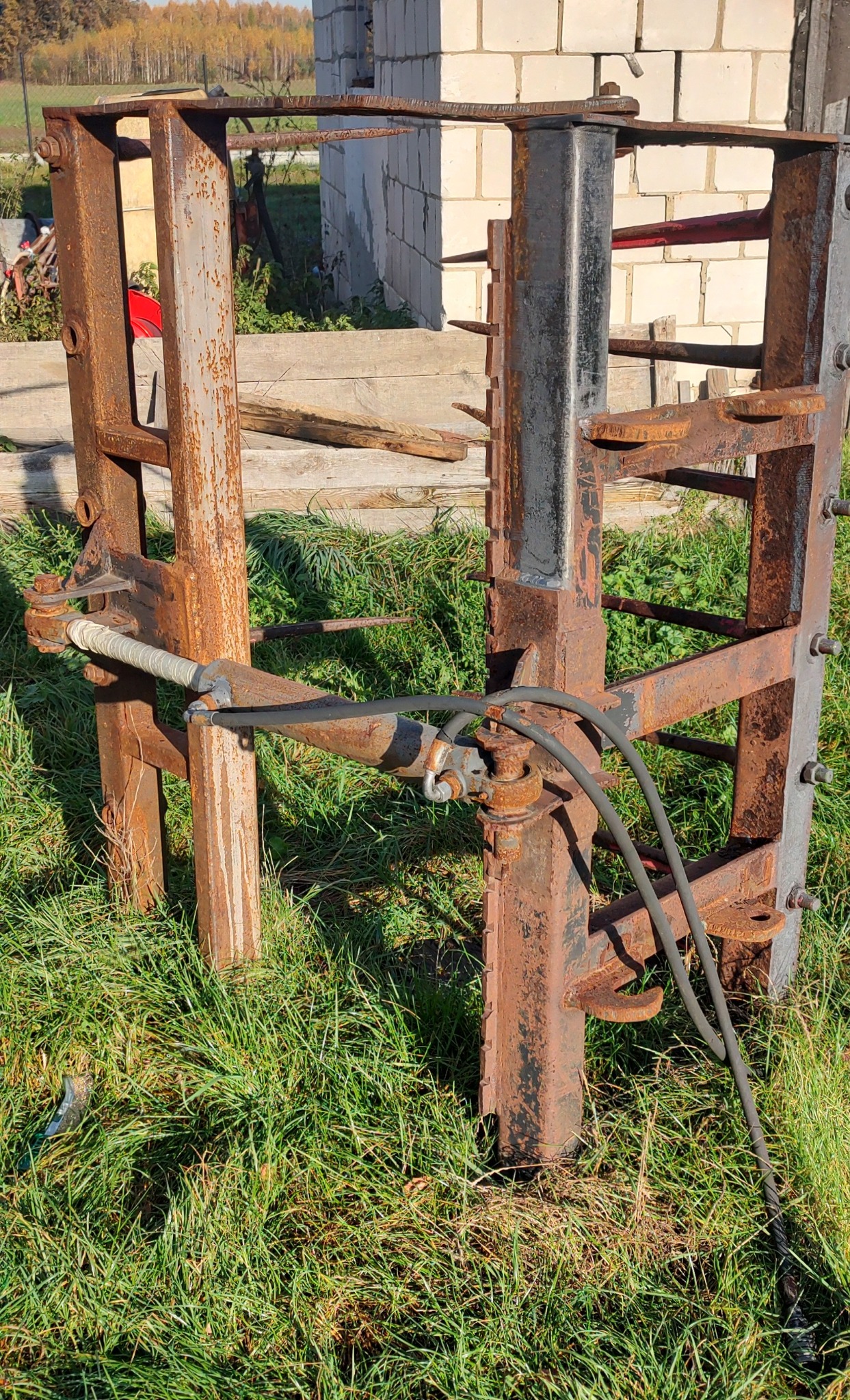 Zardzewiała, metalowa konstrukcja, prawdopodobnie element maszyny do piaskowania, stojąca na trawie przed budynkiem z białej cegły.