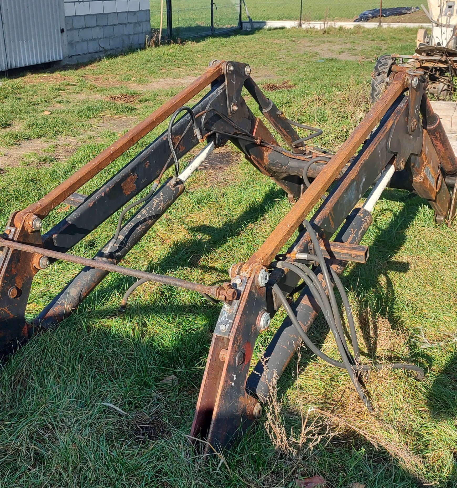 Metalowa konstrukcja ładowacza czołowego z widocznymi śladami rdzy, leżąca na trawie przed budynkiem gospodarczym.