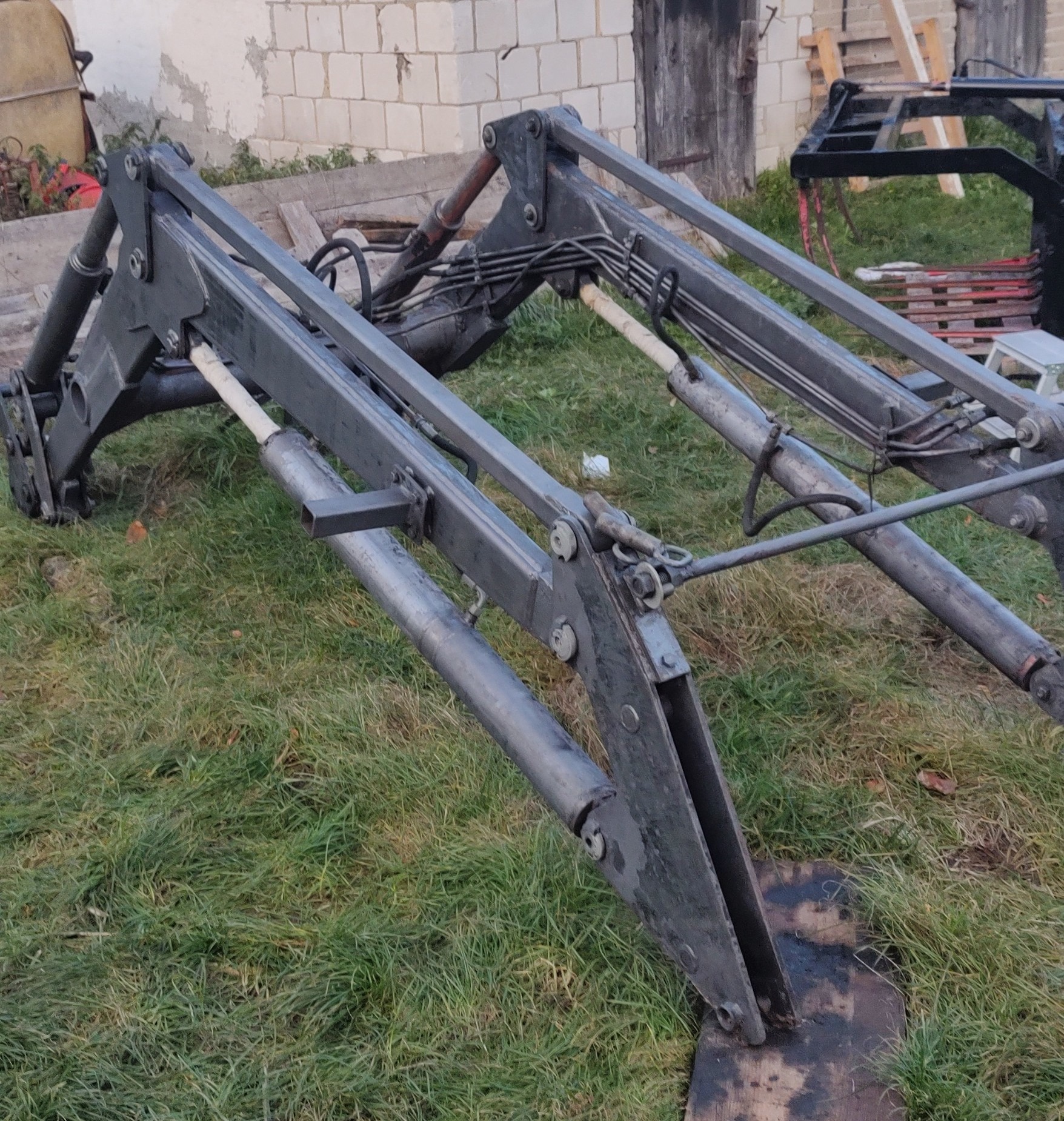 Metalowa konstrukcja ładowacza czołowego z widocznymi siłownikami hydraulicznymi, leżąca na trawie przed budynkiem z cegły i drewnianymi drzwiami.