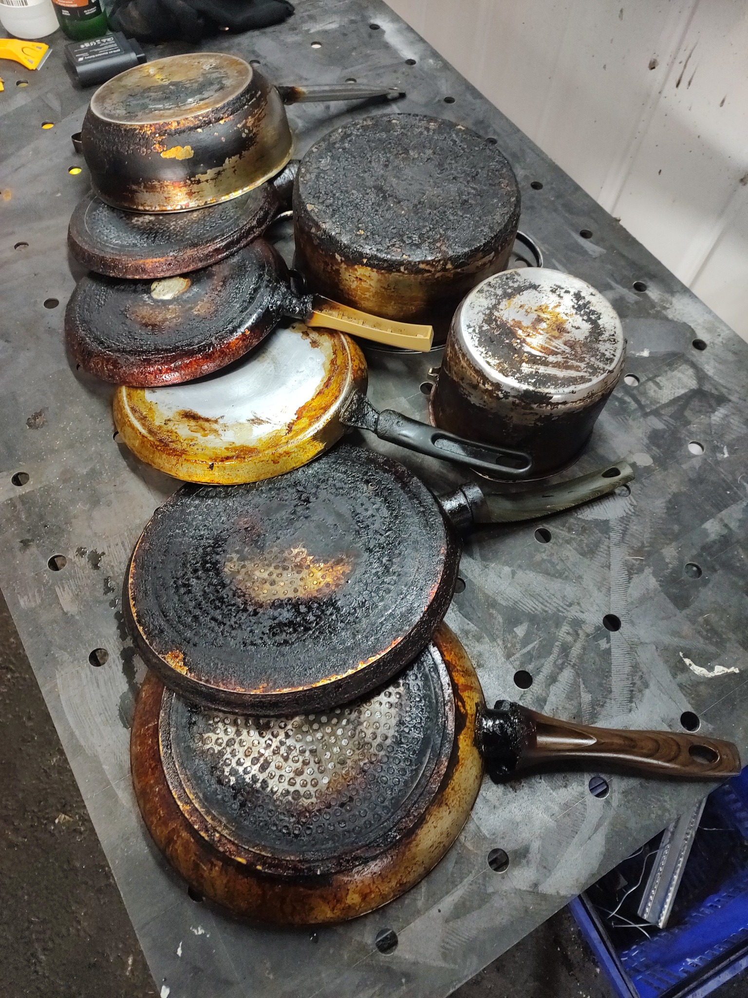 Stos mocno przypalonych garnków i patelni z resztkami zwęglonego jedzenia, leżących na metalowym stole z otworami, wymagających dokładnego wyczyszczenia.