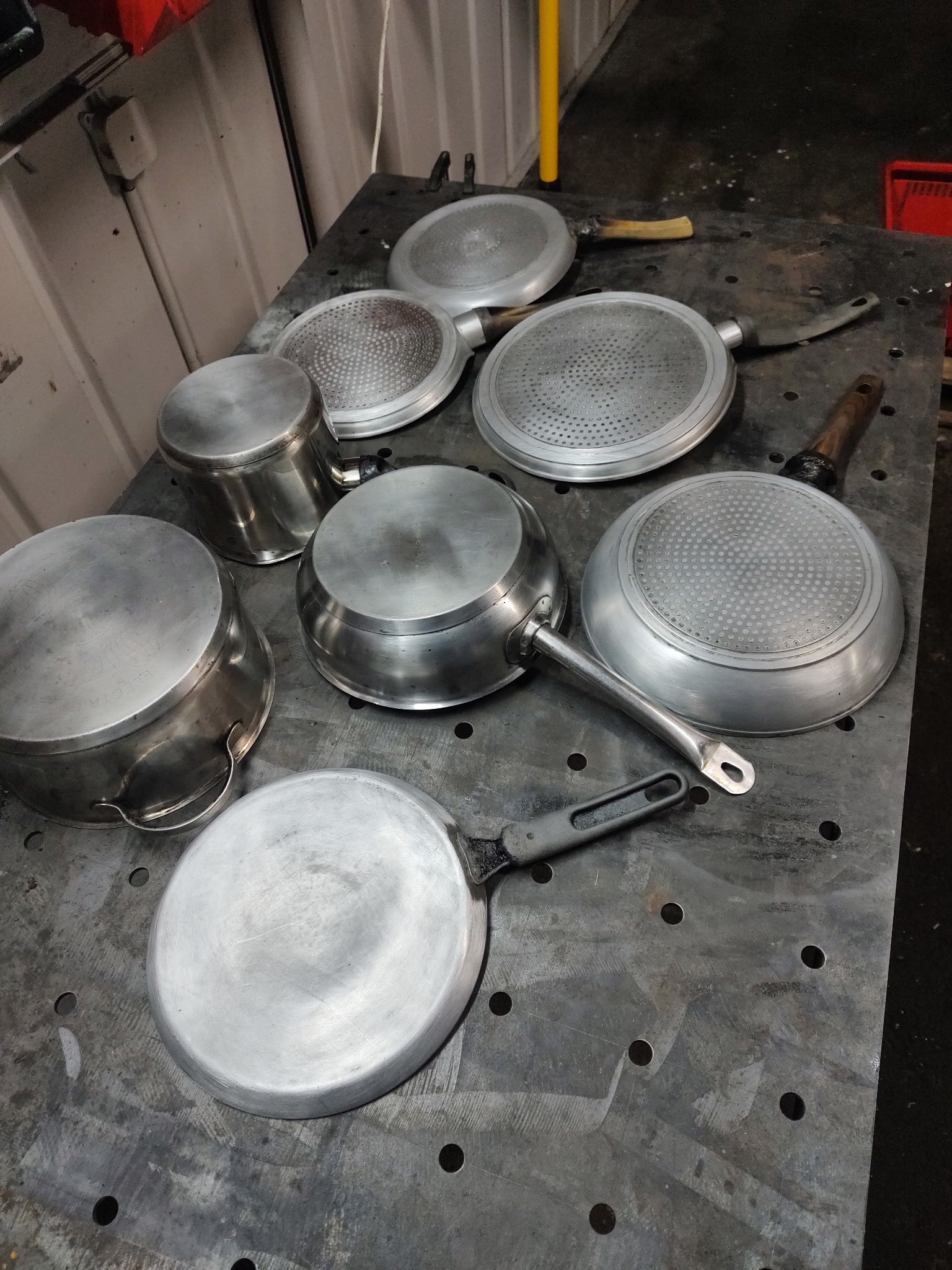 Metalowe garnki i patelnie po procesie piaskowania, leżące na perforowanym stole warsztatowym, widoczne ślady obróbki.