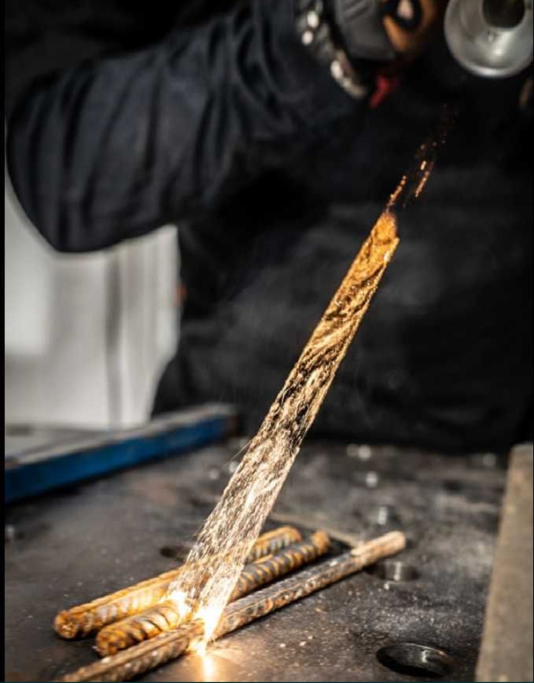 Operator w czarnym kombinezonie czyści metalowy pręt zardzewiałej stali za pomocą strumienia pod wysokim ciśnieniem, iskry i odpryski wylatują z obrabianego elementu.