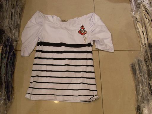 Biała bluzka w czarne paski z krótkimi bufiastymi rękawami i aplikacją kotwicy z czerwoną kokardą, leżąca na podłodze obok spakowanych ubrań.