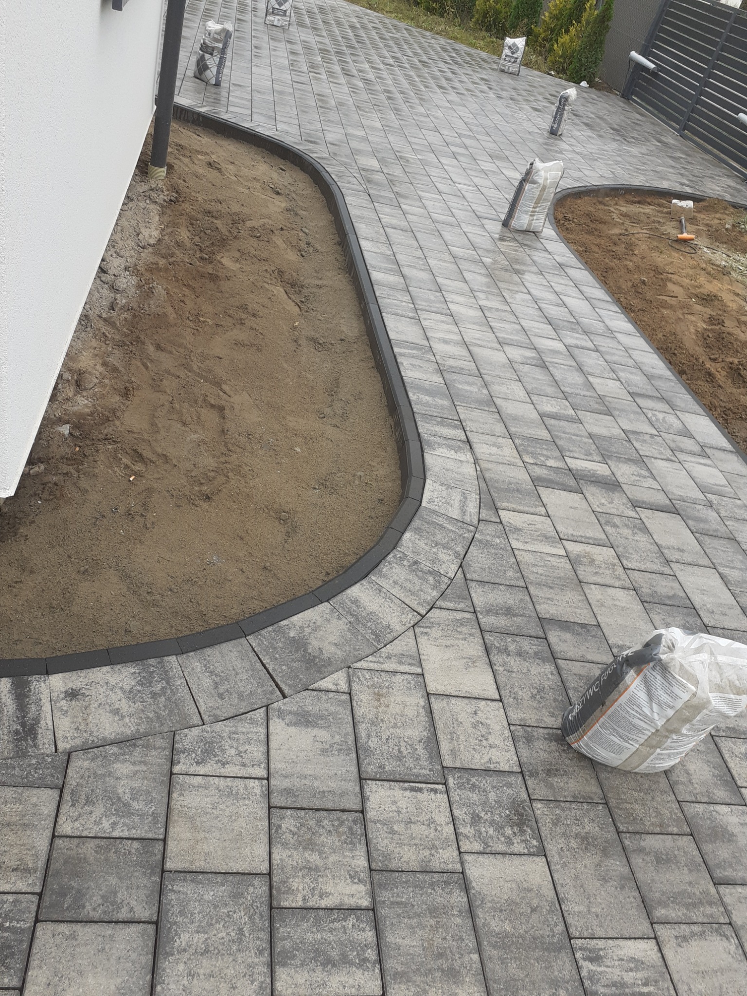 Ułożona ścieżka z kostki brukowej z obrzeżami, widoczne worki z materiałem do fugowania i przygotowane podłoże z piasku.