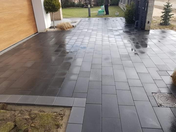 Świeżo ułożona, ciemnoszara kostka brukowa przed nowoczesnym domem z drewnianą bramą garażową, widoczny właz kanalizacyjny po prawej stronie, trawnik i drzewka w tle.