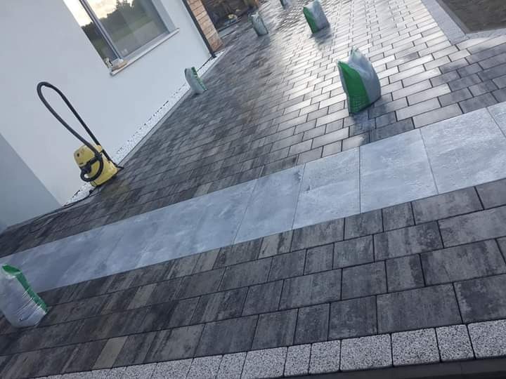 Ułożona kostka brukowa w odcieniach szarości z centralnym pasem z płyt, uzupełniana białym grysem przy ścianie domu, z workami z materiałem i odkurzaczem widocznymi na powierzchni.