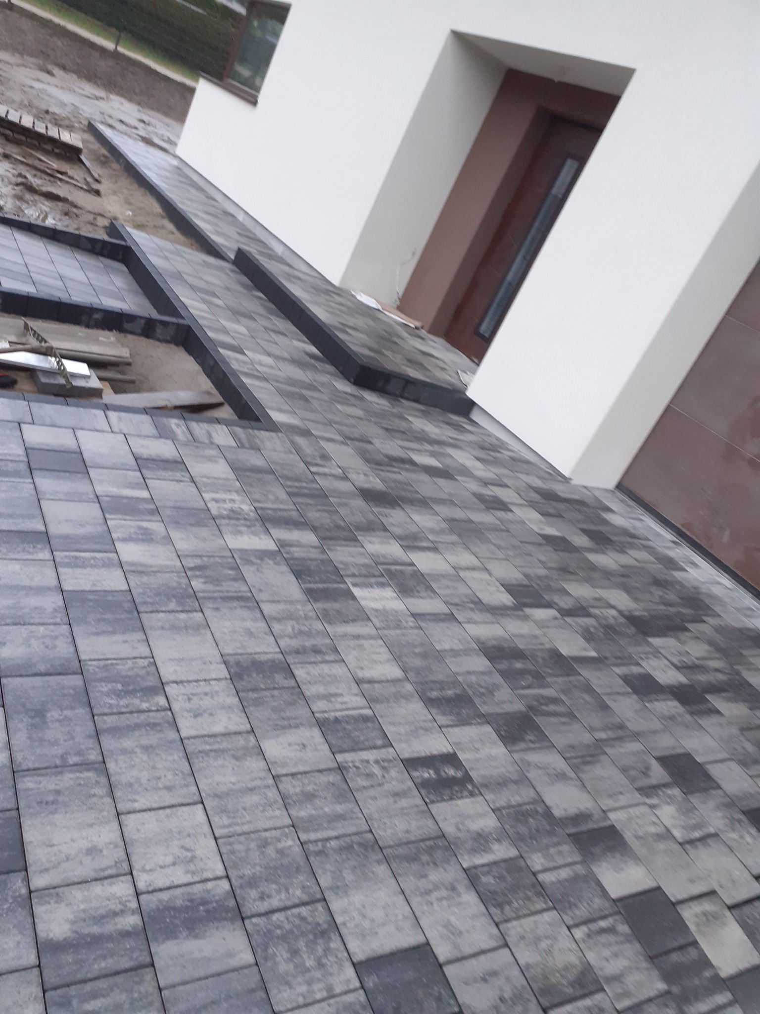 Fragment posesji w trakcie prac brukarskich, widoczne ułożone szare kostki brukowe oraz przygotowany teren pod dalsze prace, na drugim planie fragment budynku z oknem i drzwiami.