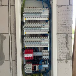 Rozdzielnica elektryczna w białej obudowie, zamontowana na ścianie z betonu komórkowego z wypisanymi nazwami obwodów. Widoczne przewody i zabezpieczenia.