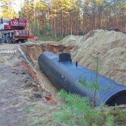 ADONIS Ogrody Andrzej Judziński - Czarny zbiornik zakopany w ziemi, widoczny wykop, piasek i żuraw budowlany w tle, sugerujący instalację podziemnego systemu.