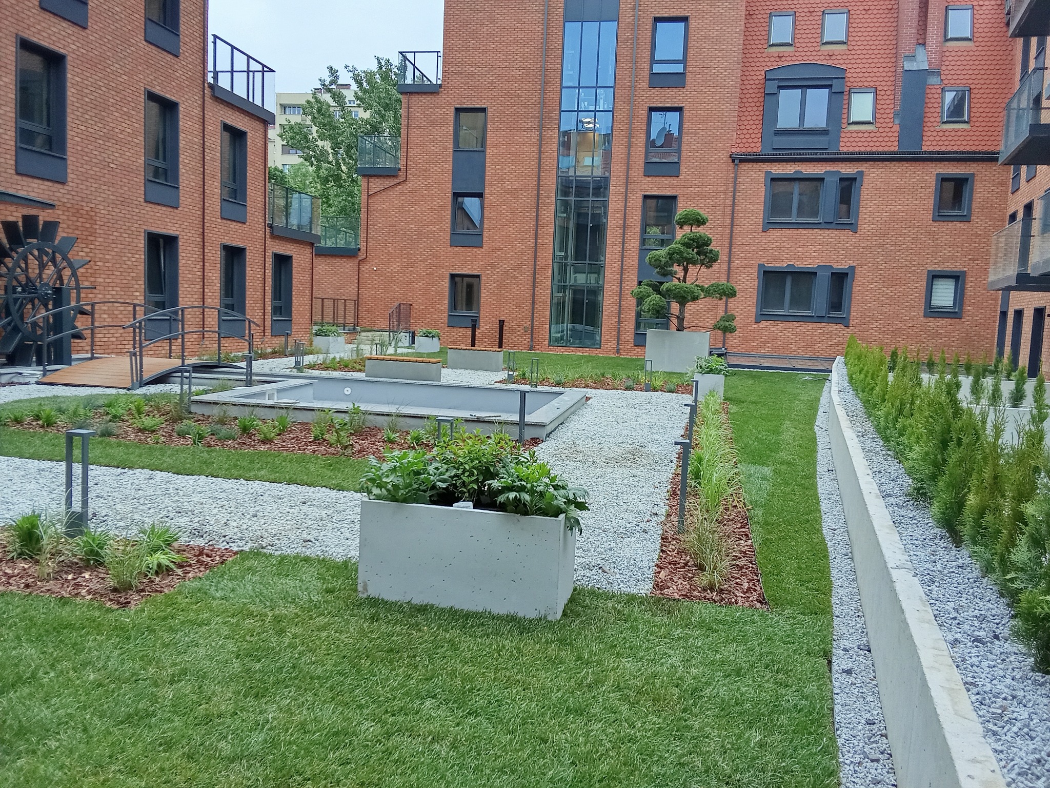 Nowoczesny ogród miejski z zielonym trawnikiem, ścieżkami z białego żwiru, betonowymi donicami z roślinami i fontanną na tle ceglanej architektury.