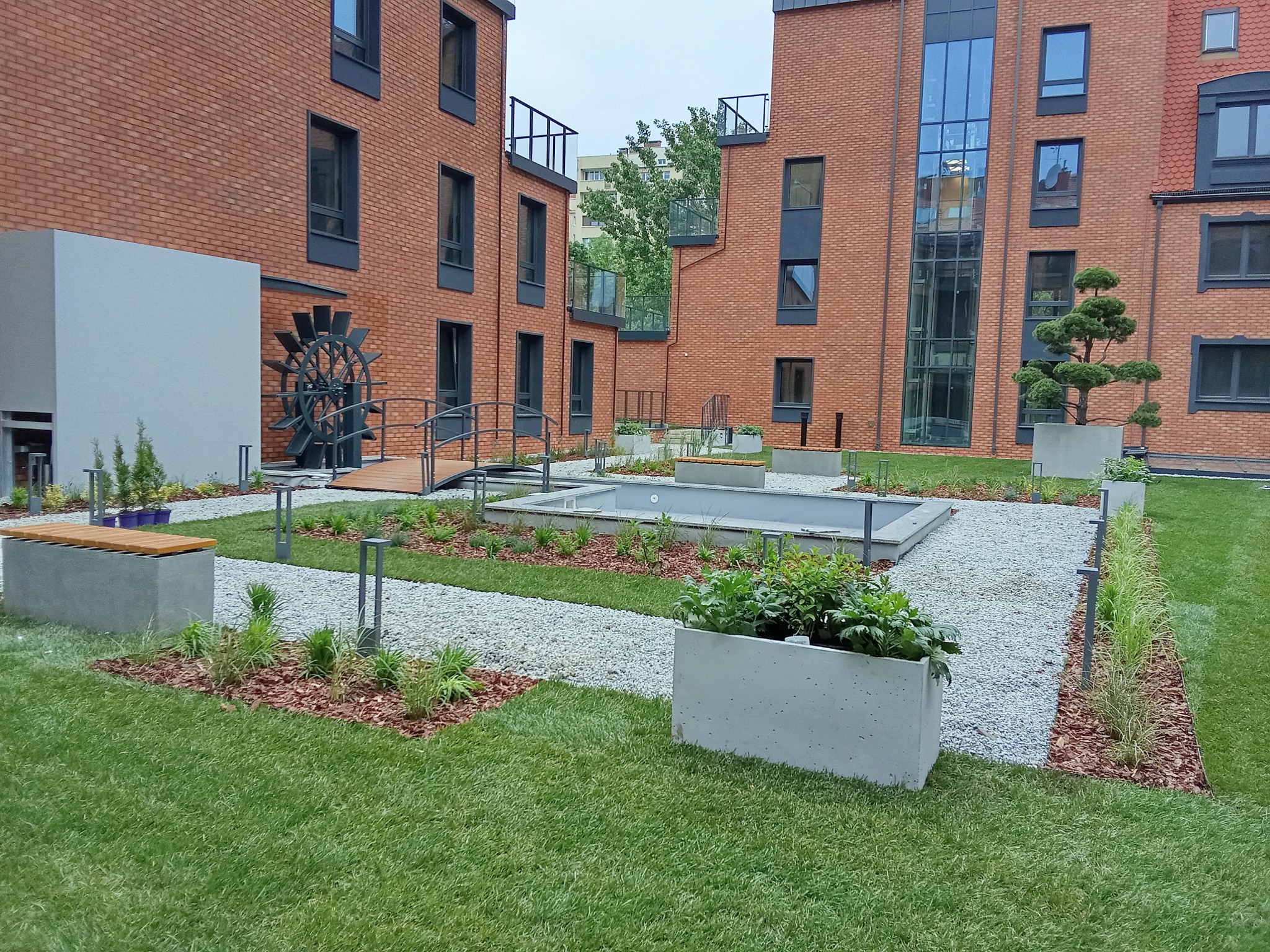 Nowoczesny ogród z elementami wodnymi i dekoracyjnym młynem wodnym, otoczony budynkami z czerwonej cegły. Zadbany trawnik, ścieżki z jasnego żwiru.