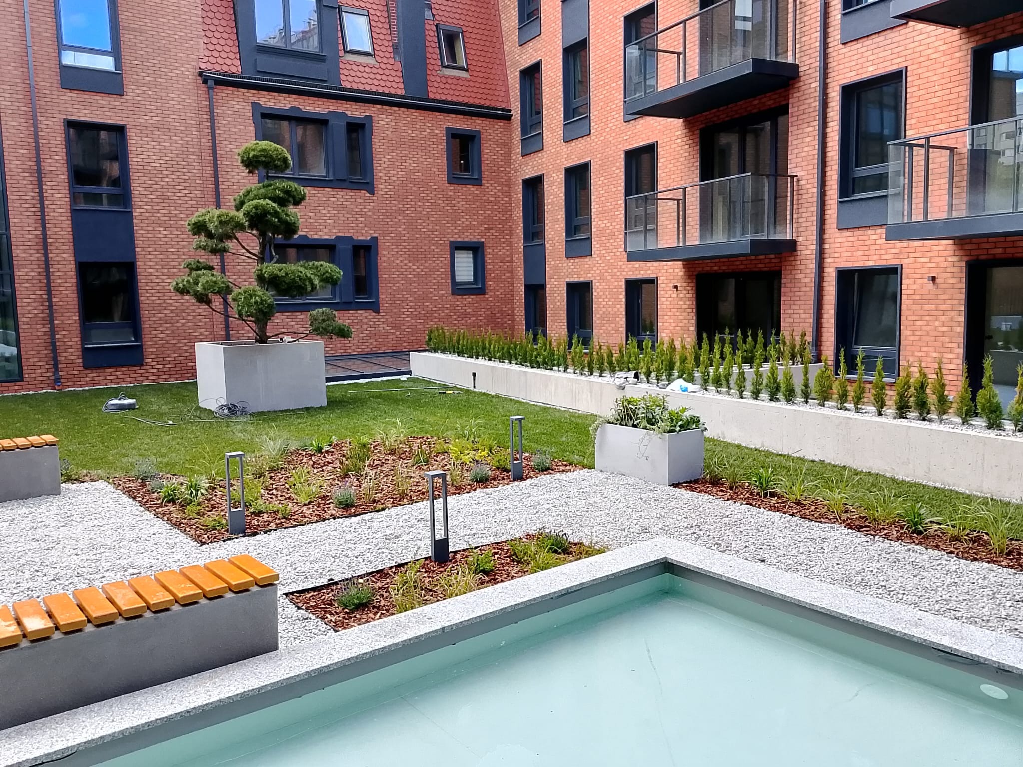 Estetyczny dziedziniec z zielenią i elementami małej architektury, w tle budynek z czerwonej cegły. W centralnej części trawnik, ścieżki z białego żwiru i niskie rośliny.