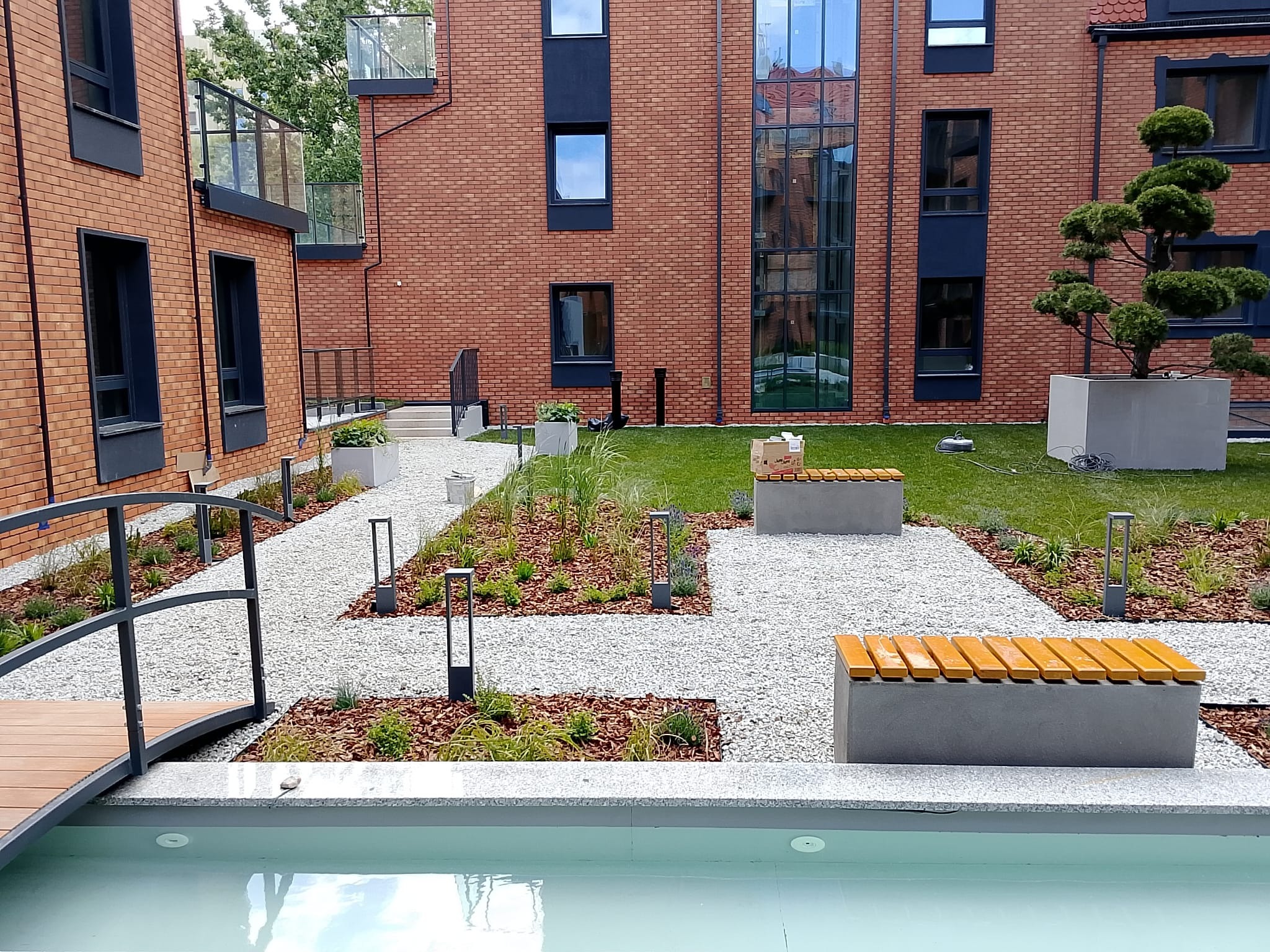 Nowoczesny ogród wewnątrz osiedla: ścieżki z białego żwiru, drewniany mostek nad płytkim basenem, minimalistyczne ławki i starannie przycięte drzewo.