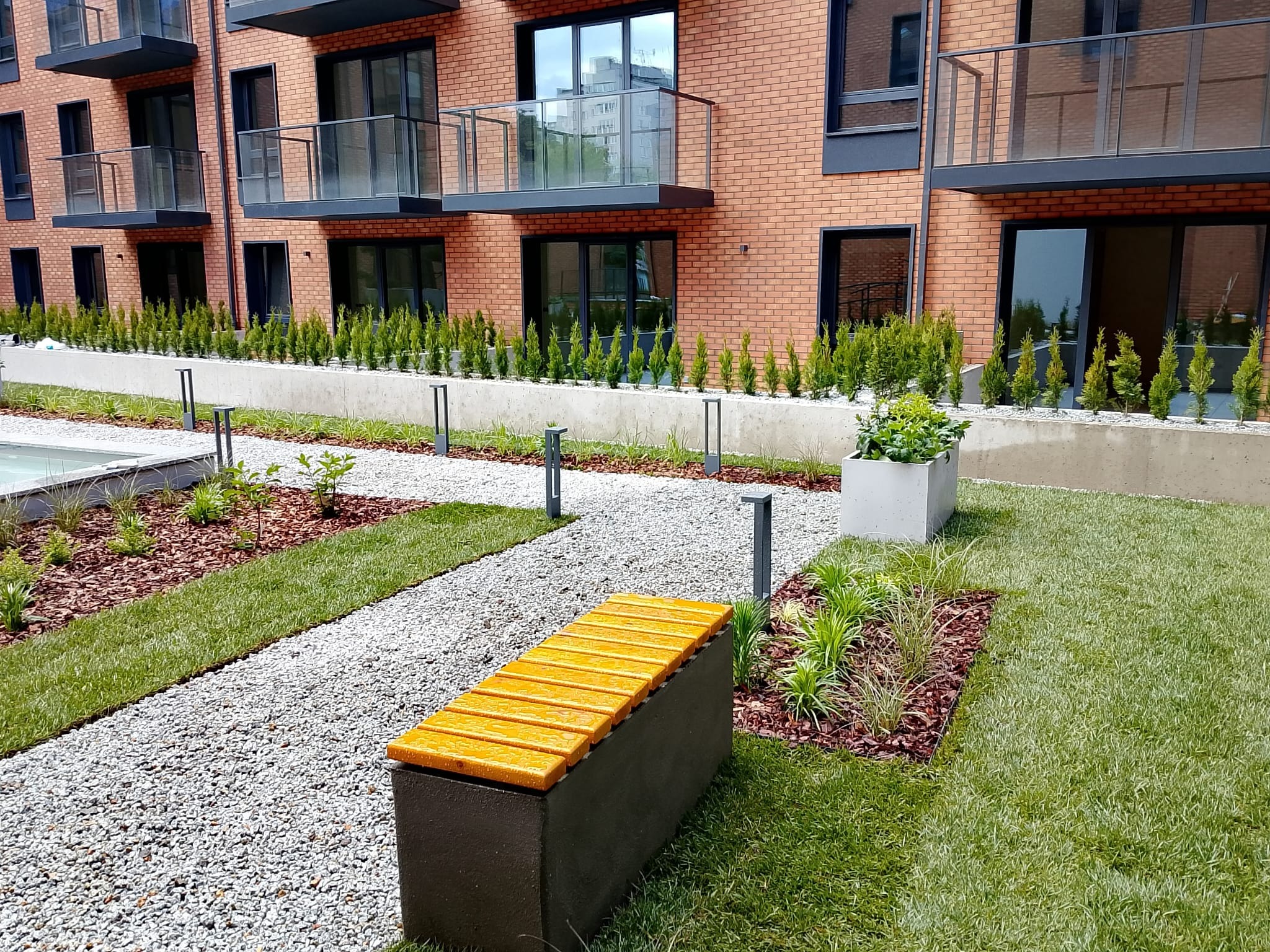 Nowoczesny ogród przy apartamentowcu: żółta ławka na żwirowej ścieżce, rabaty z korą i roślinami, trawnik, rząd tuj i ceglany budynek z balkonami.