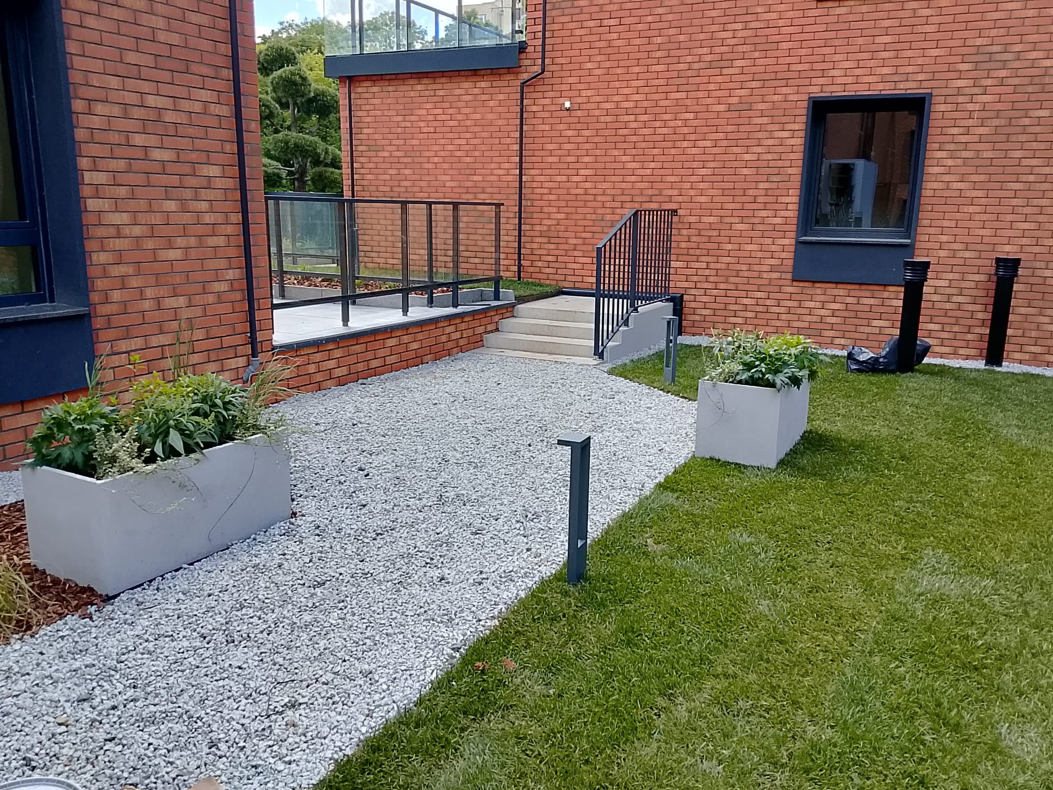 Ogród przy nowoczesnym budynku w Lubinie: ścieżka z białego żwiru, donice z zielenią, trawnik i elementy oświetlenia. Architektura ogrodowa.