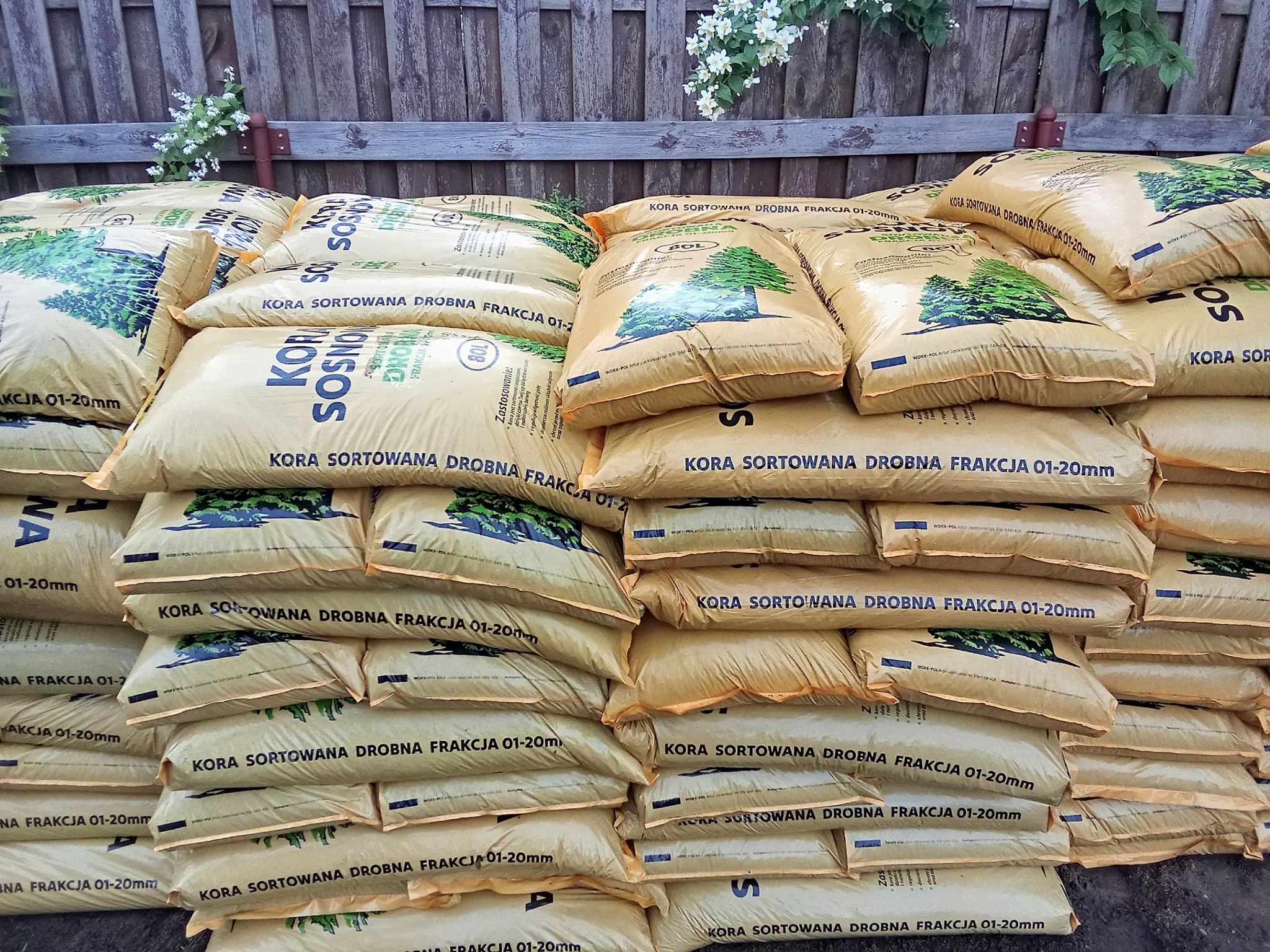 Stos worków z korą sosnową sortowaną, drobna frakcja 01-20mm, ułożonych na zewnątrz przy drewnianym płocie. Widoczne kwiaty i zieleń w tle.