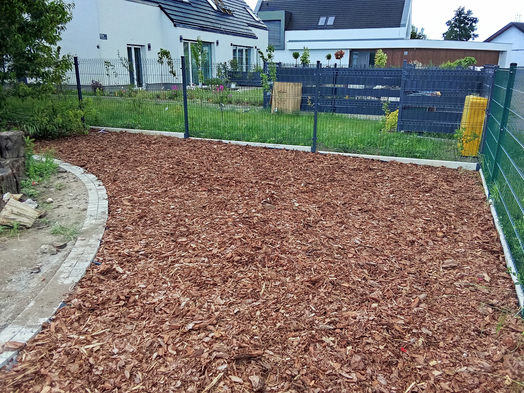 Ściółkowanie kory w ogrodzie przydomowym, widok z góry. Kora równomiernie rozłożona, obrzeże z kostki, w tle nowoczesny dom i metalowe ogrodzenie.