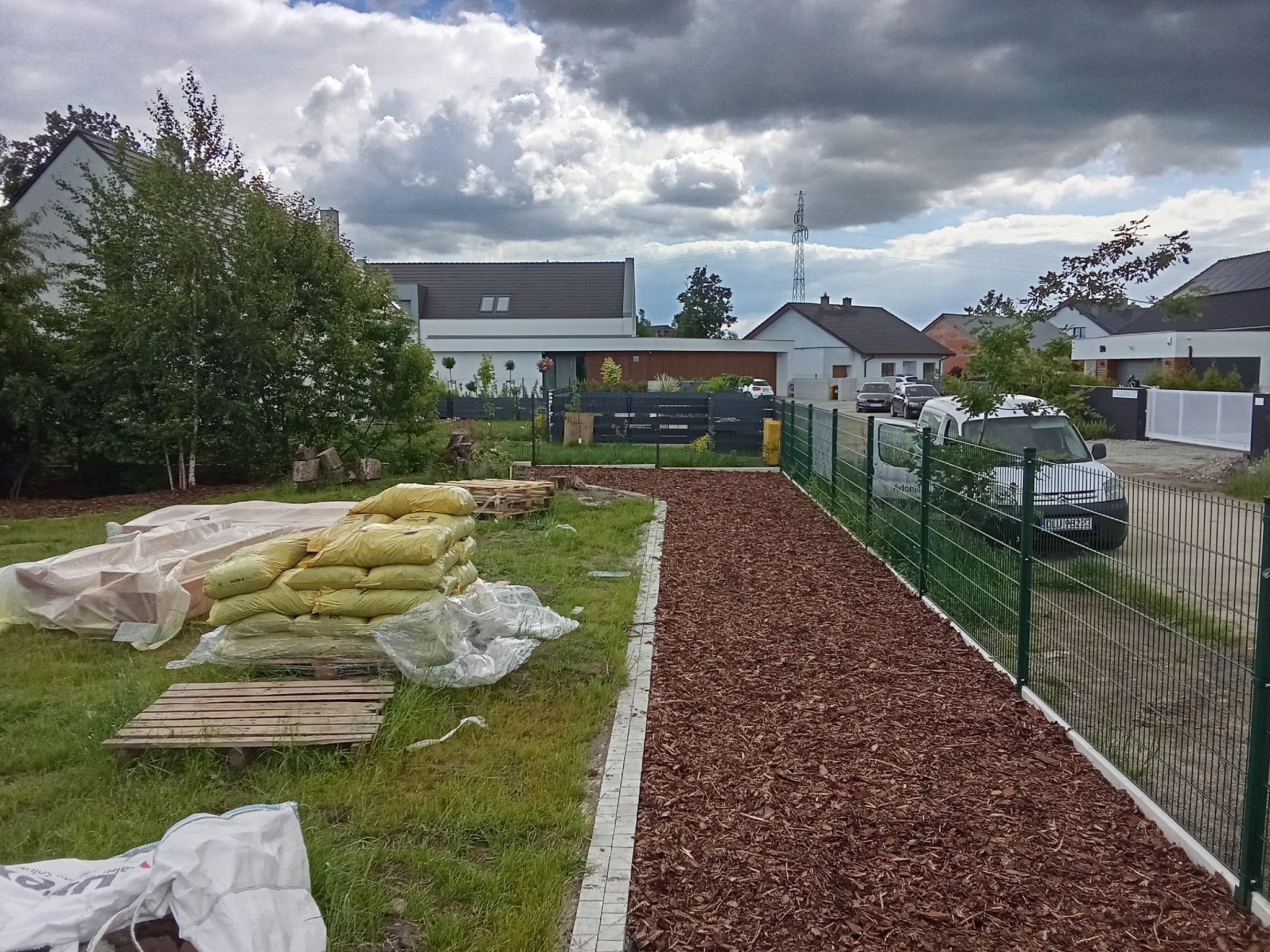 Przygotowany teren z brązową korą i obrzeżem z kostki, worki z materiałem budowlanym i ogrodzeniem panelowym w tle, prawdopodobnie przed założeniem ogrodu.