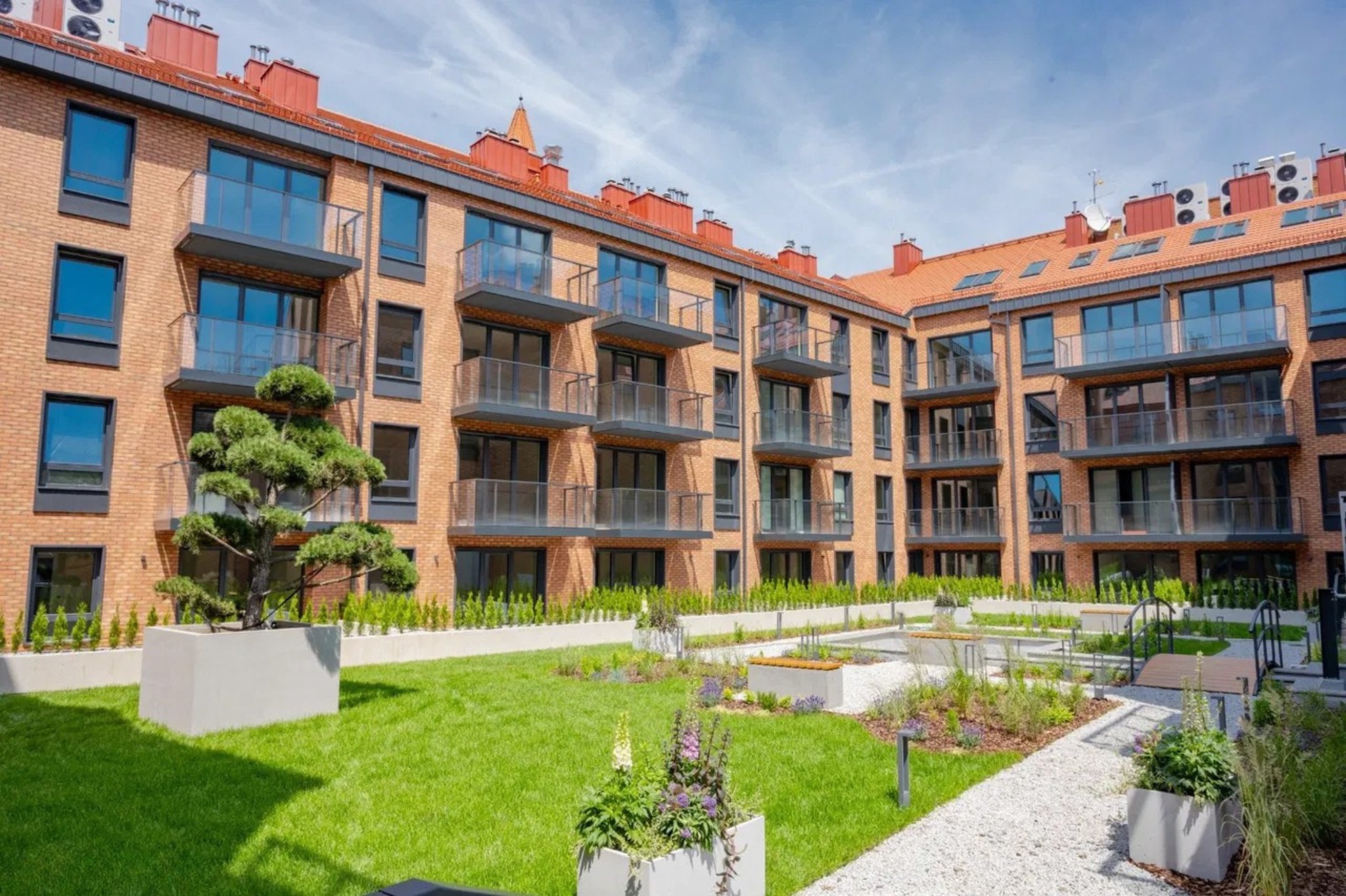 Dziedziniec nowoczesnego apartamentowca z zielonym trawnikiem, zadbaną roślinnością w donicach i białym żwirem. Budynek z cegły z balkonami w tle.