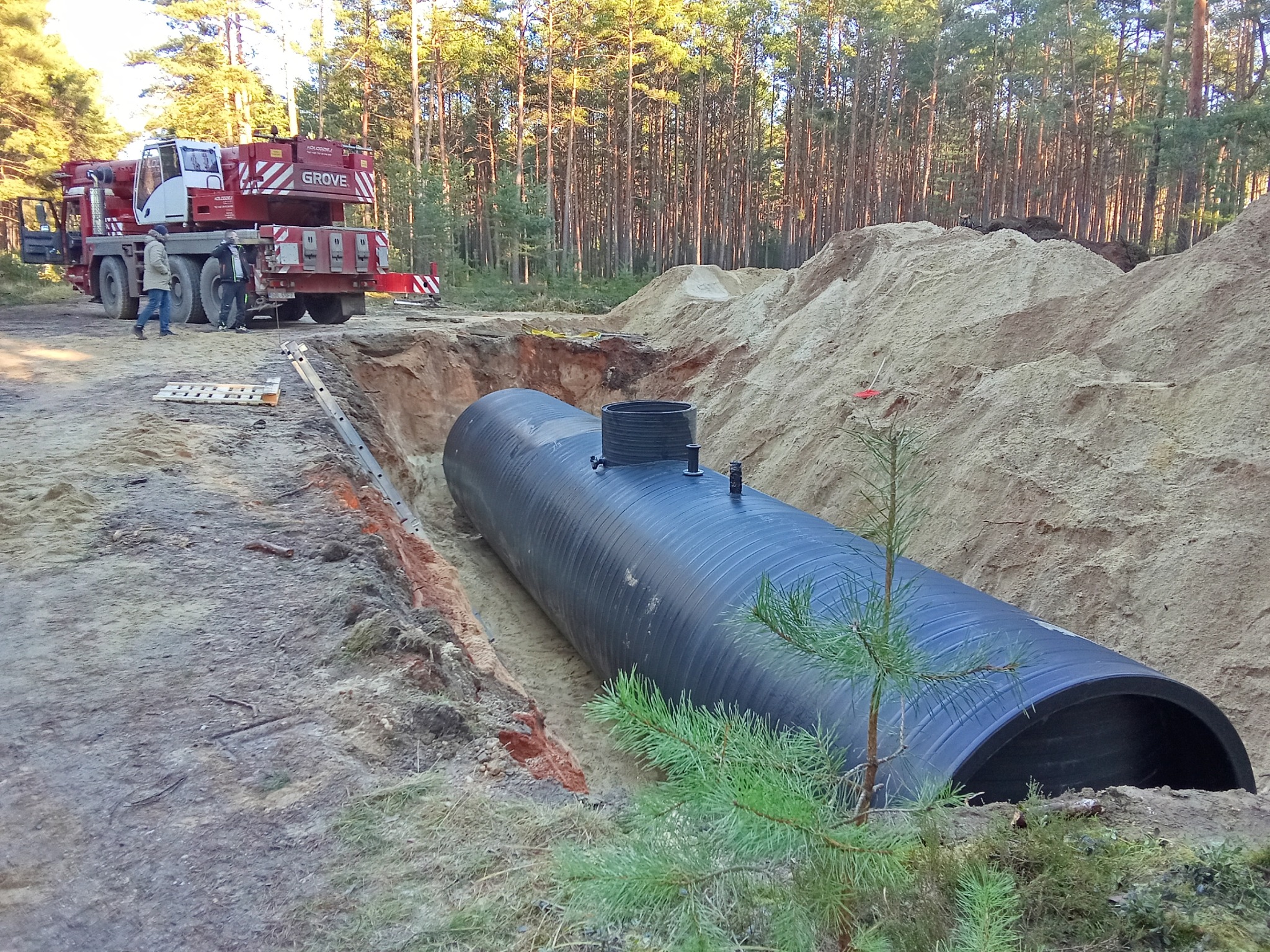 Montaż podziemnego zbiornika na wodę w Lubinie. Widoczny czarny zbiornik w wykopie, obok kopiec piasku i dźwig. Prace ziemne w toku, las w tle.