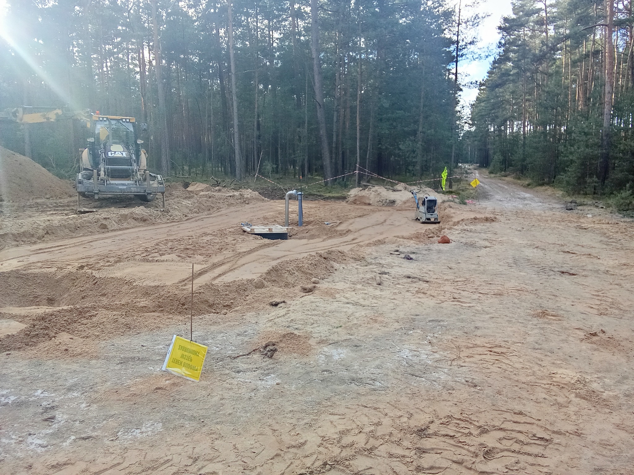 Przygotowanie terenu pod budowę w lesie: ciężki sprzęt CAT, ubijarka, wykopane rury, żółta tabliczka ostrzegawcza i pracownik w odblaskowej kamizelce.