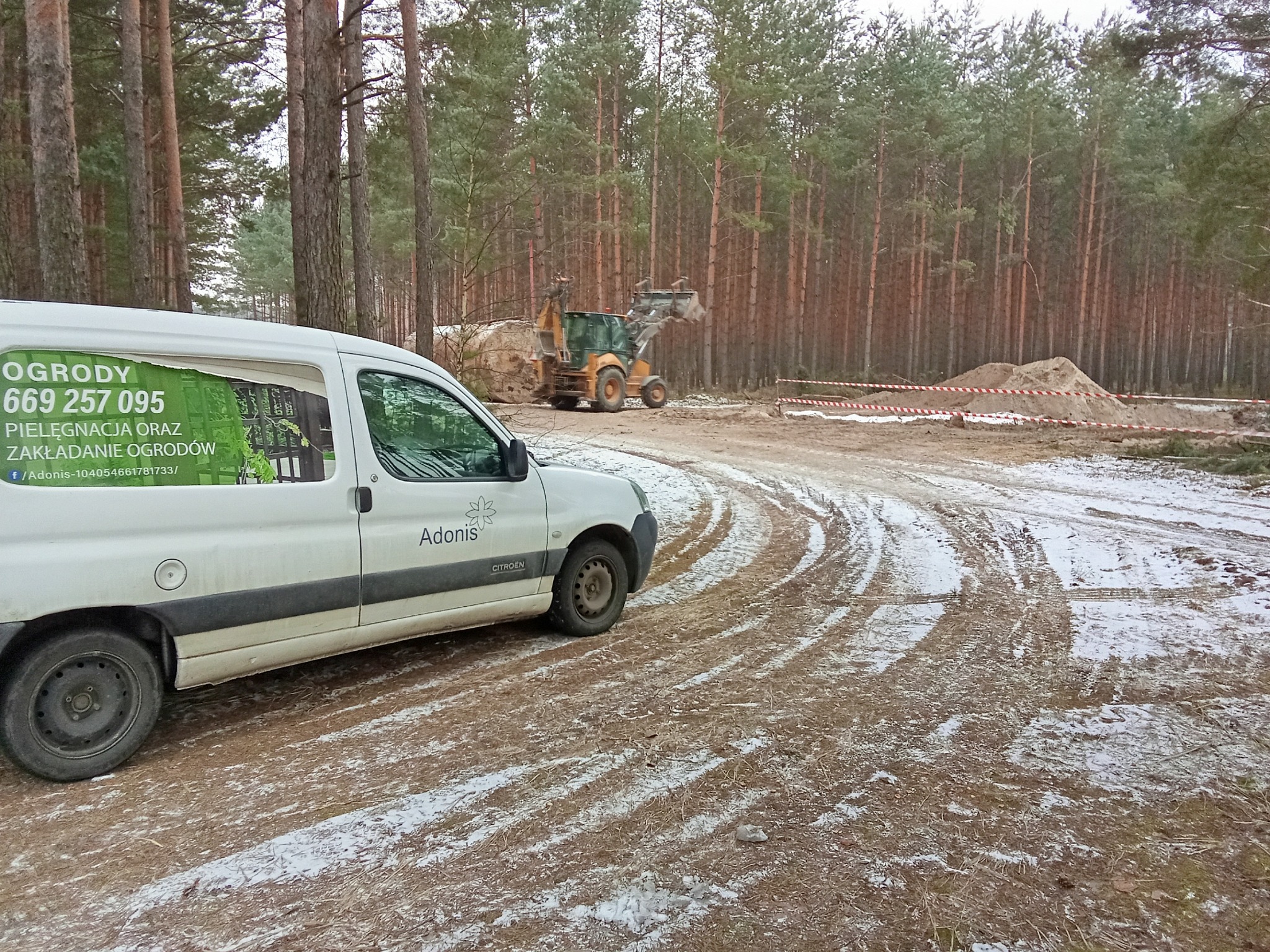 Biały van firmy ogrodniczej Adonis na tle lasu i sprzętu budowlanego. Na pojeździe widoczne dane kontaktowe i zakres usług: pielęgnacja i zakładanie ogrodów.