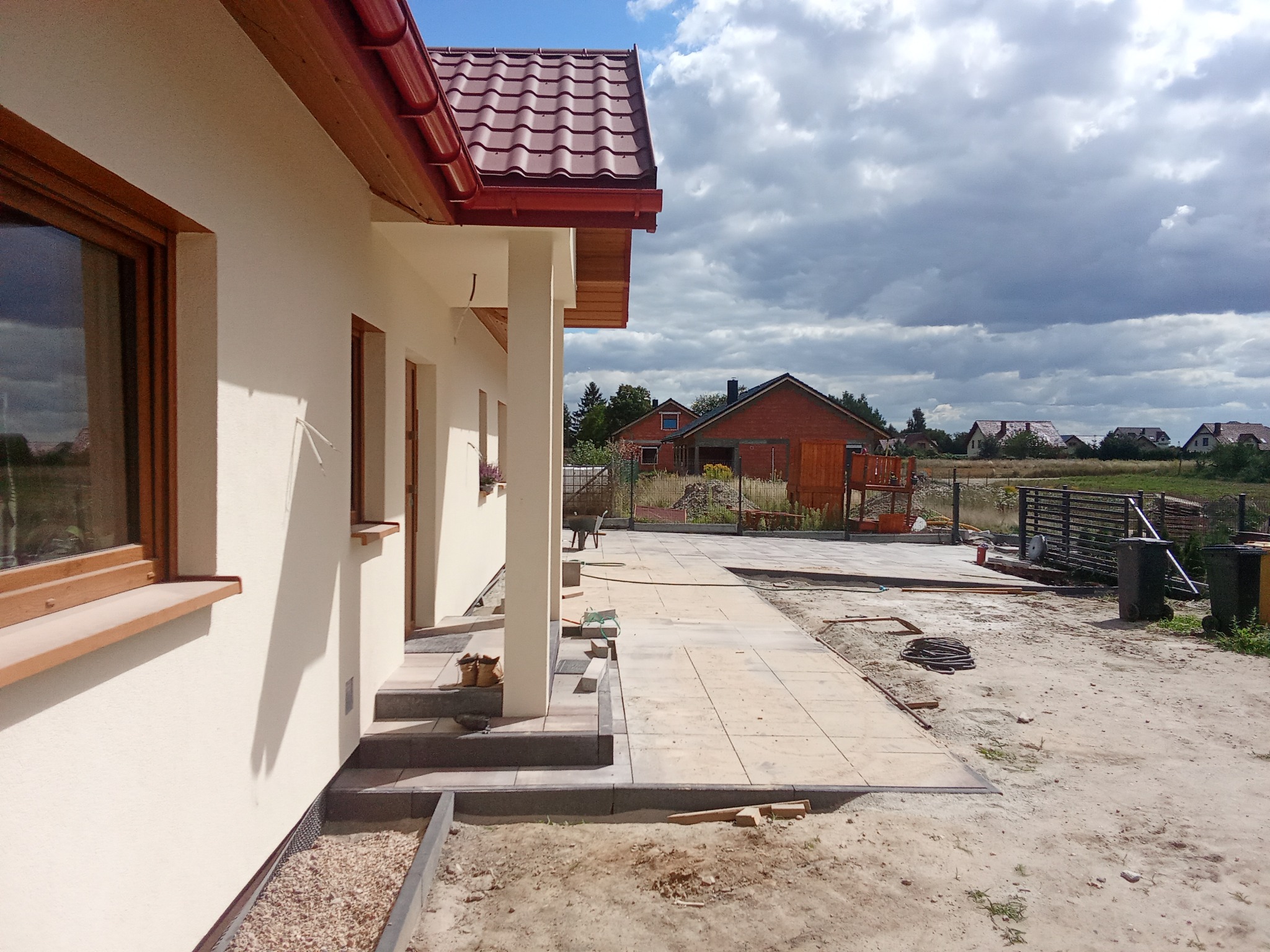 Jasna elewacja domu z brązowymi oknami i czerwonym dachem. Ułożona kostka brukowa tworzy ścieżkę. Widoczne obrzeża i przygotowane podłoże. W tle zabudowa jednorodzinna.