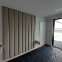 Beżowy panel tapicerowany pionowymi pasami na ścianie w sypialni, obok biała szafka nocna i drzwi balkonowe, podłoga zabezpieczona folią malarską.